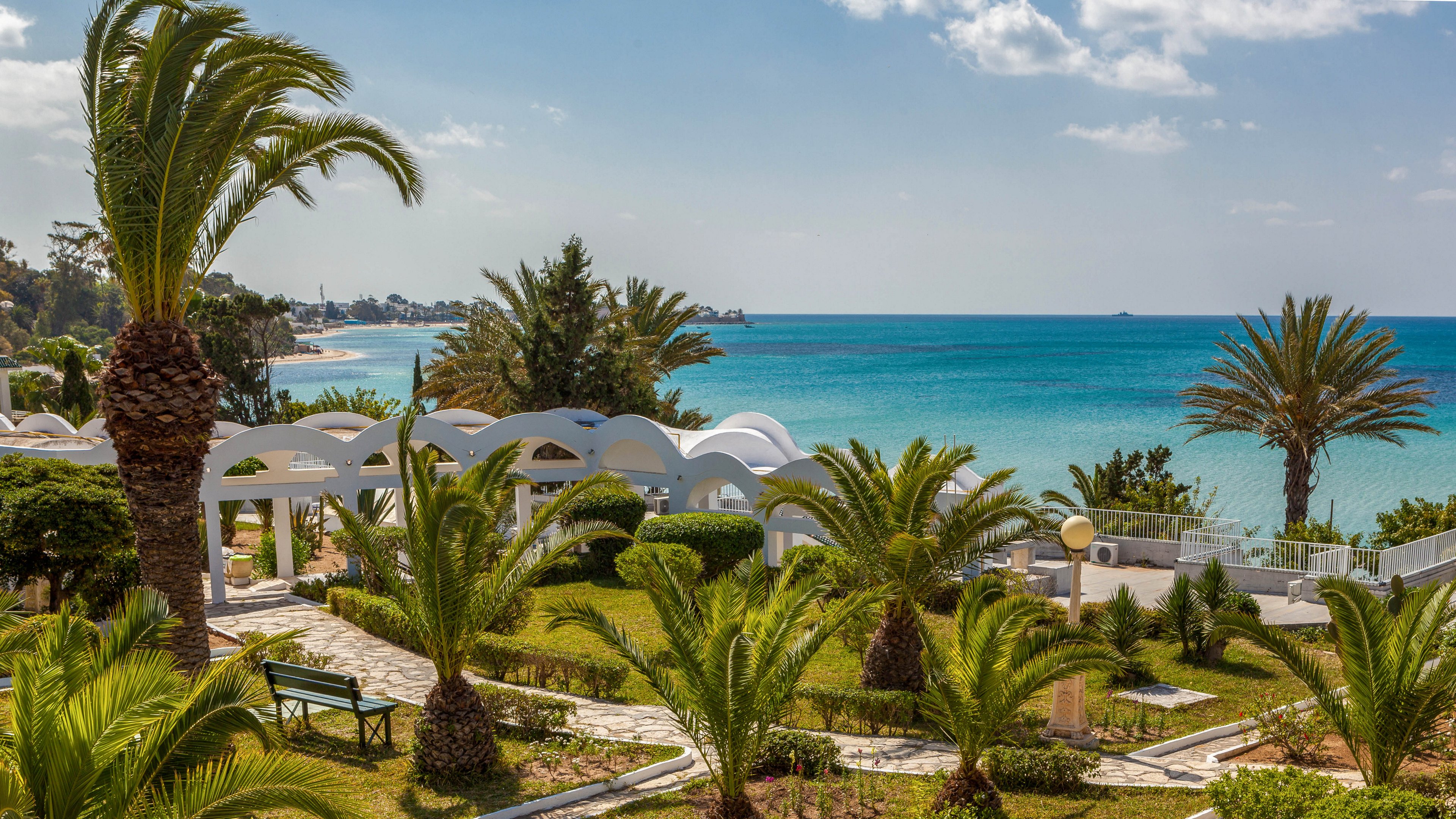 Lush palm trees and manicured gardens frame a view of a calm turquoise sea. White arched structures and pathways create a resort-like atmosphere along the coastline. The sky is mostly clear with a few scattered clouds, enhancing the bright and sunny mood. The scene suggests a peaceful coastal retreat ideal for relaxation and leisure.