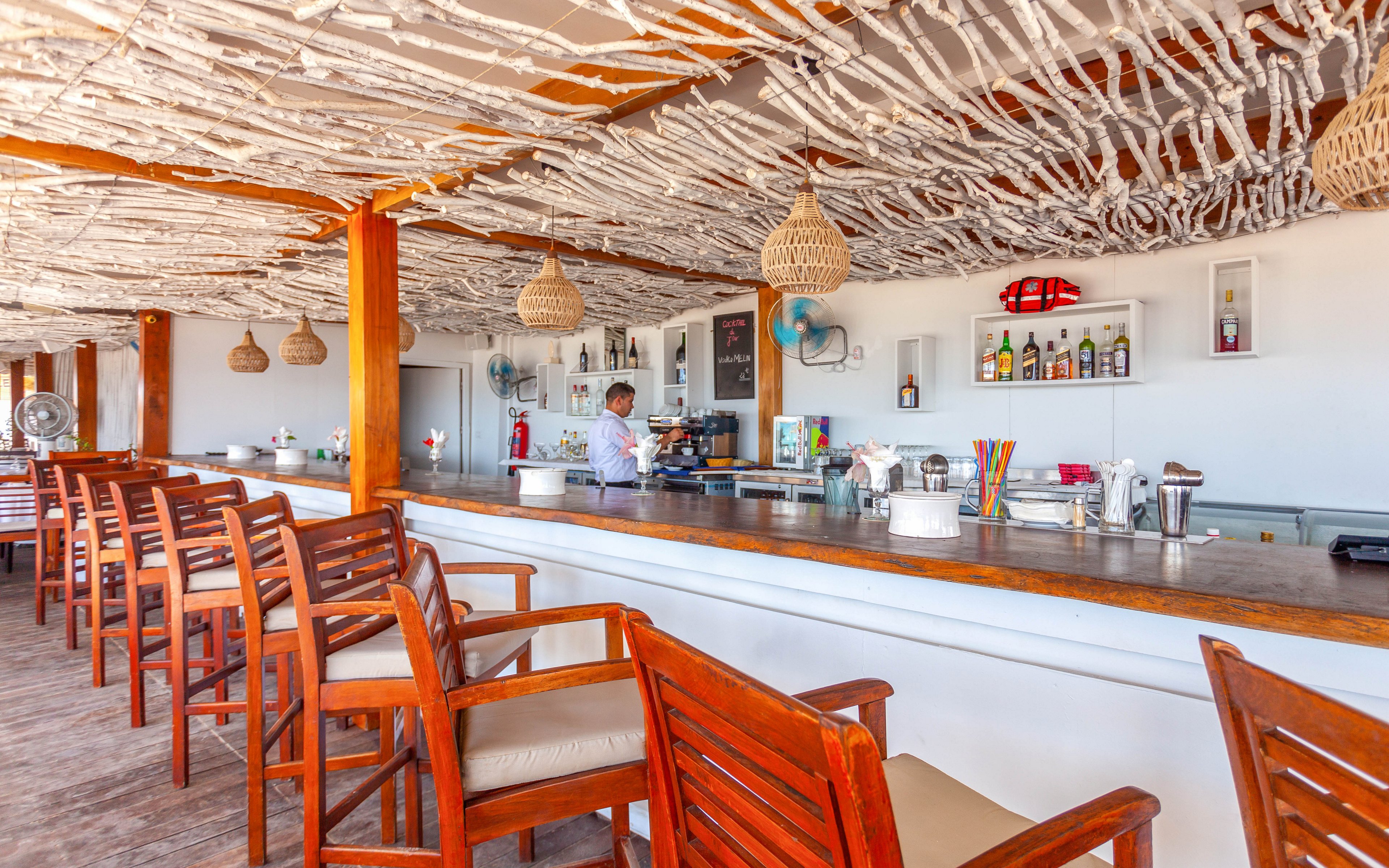 An open-air beach bar features a long wooden counter lined with high wooden chairs. A bartender works behind the bar, surrounded by bottles, glassware, and colorful straws. The ceiling is decorated with white driftwood-style branches and woven pendant lights, creating a coastal atmosphere. Sunlight fills the space, emphasizing the relaxed, tropical setting and casual vacation vibe.