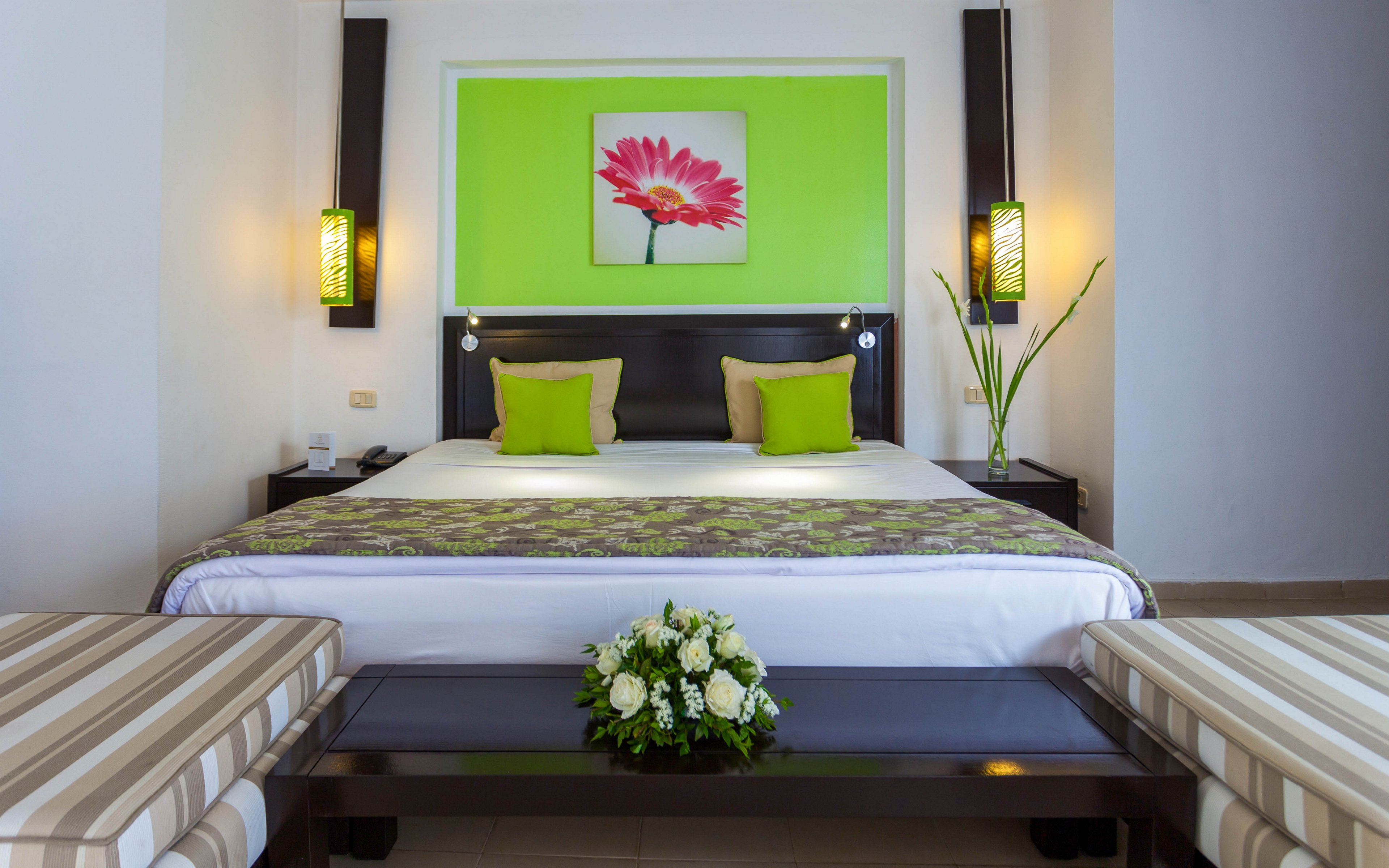 A neatly made double bed is centered in a modern hotel room with bright green accents. Two striped benches sit at the foot of the bed, framing a low black table with a white floral arrangement. Matching green pillows and a patterned runner coordinate with the green wall panel and floral artwork above the headboard. Soft wall sconces provide warm lighting, creating a clean and inviting atmosphere.