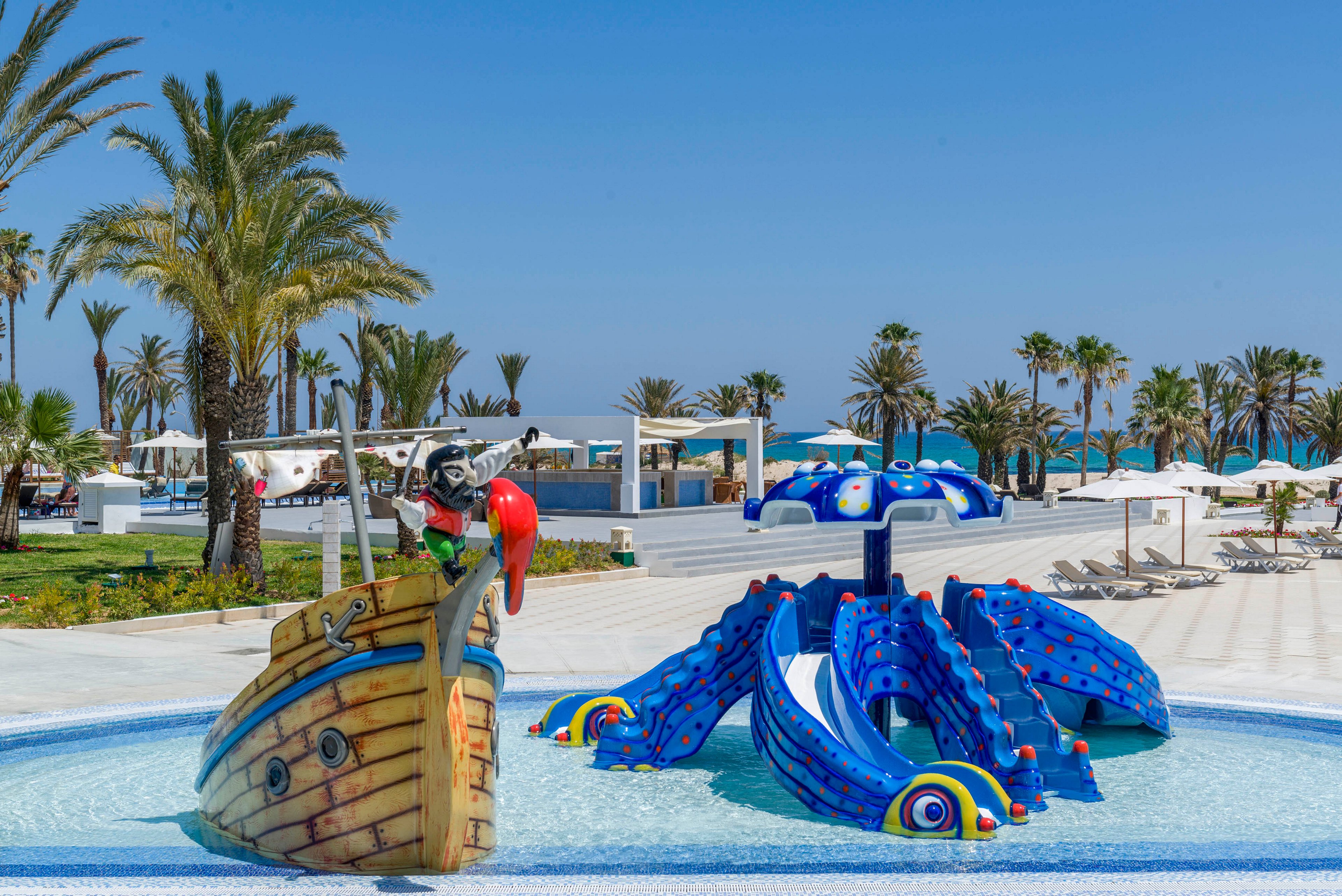 A vibrant children’s water play area features a pirate ship structure and a blue octopus slide in a shallow pool. The scene is set at a sunny resort with palm trees, white loungers, and umbrellas in the background. Bright primary colors and clear blue skies create a cheerful, tropical atmosphere. The area appears calm and inviting, ideal for family vacations and kids’ activities.