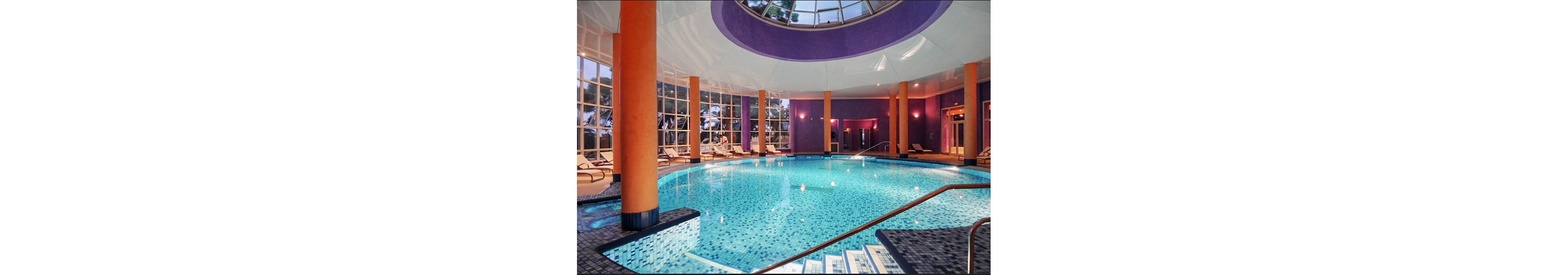 A spacious indoor swimming pool is surrounded by lounge chairs and tall orange columns. Large floor-to-ceiling windows let in natural light and reveal trees outside. The pool features blue mosaic tiles and stainless steel handrails leading into the water. Soft purple walls and warm lighting create a relaxing, resort-like atmosphere.