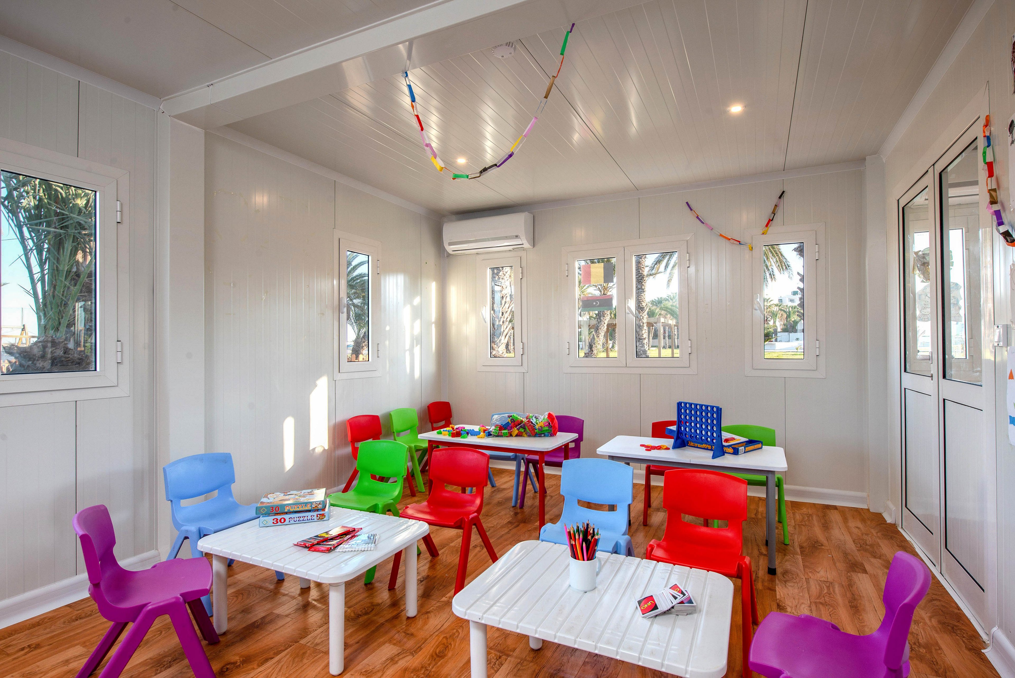 A bright classroom is furnished with small white tables and colorful plastic chairs arranged around the room. Toys, books, and art supplies are placed on the tables, suggesting a space for young children’s activities. The room has wood-style flooring, multiple windows, and an air conditioning unit mounted on the wall. Simple hanging decorations and recessed ceiling lights add to the cheerful, welcoming atmosphere.