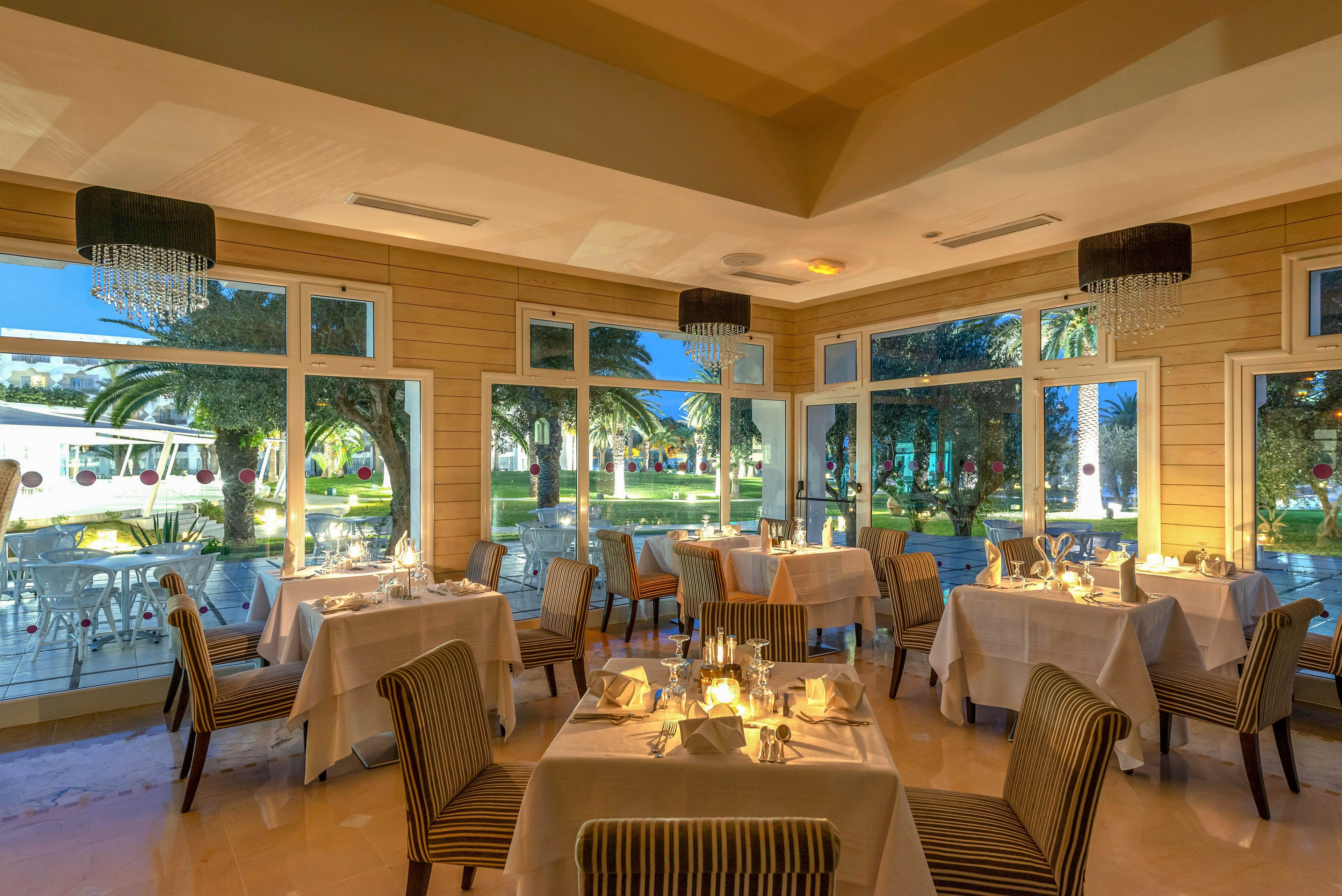Upscale restaurant dining room with neatly set tables is shown in warm evening light. Large windows reveal a lush garden with palm trees and outdoor seating. Candles and folded napkins create an intimate, romantic atmosphere. Neutral tones, soft lighting, and contemporary chandeliers contribute to a relaxed yet sophisticated setting.