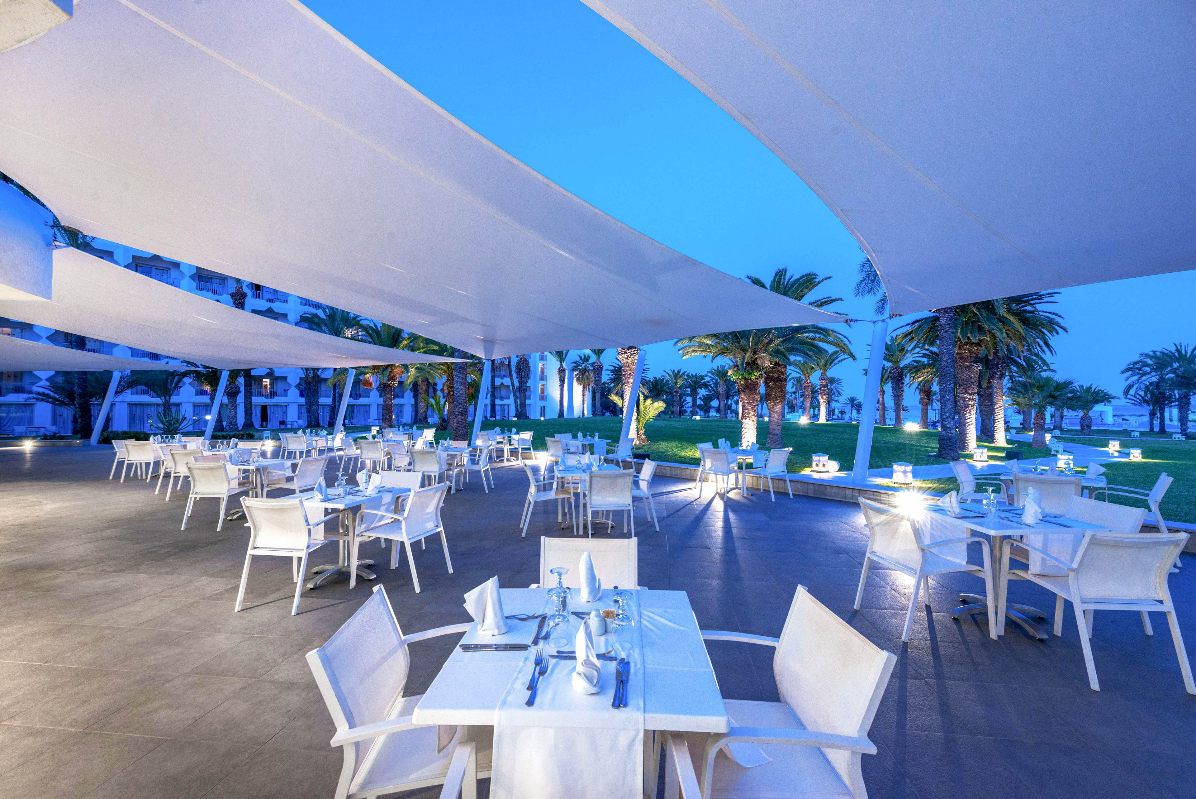An elegant open-air restaurant is set with white tables and chairs under large modern canopy sails. The scene is captured at dusk with a cool blue ambiance and soft lighting. Palm trees and manicured lawns frame the dining area, suggesting a coastal or resort setting. The overall atmosphere feels calm, spacious, and inviting for evening dining.