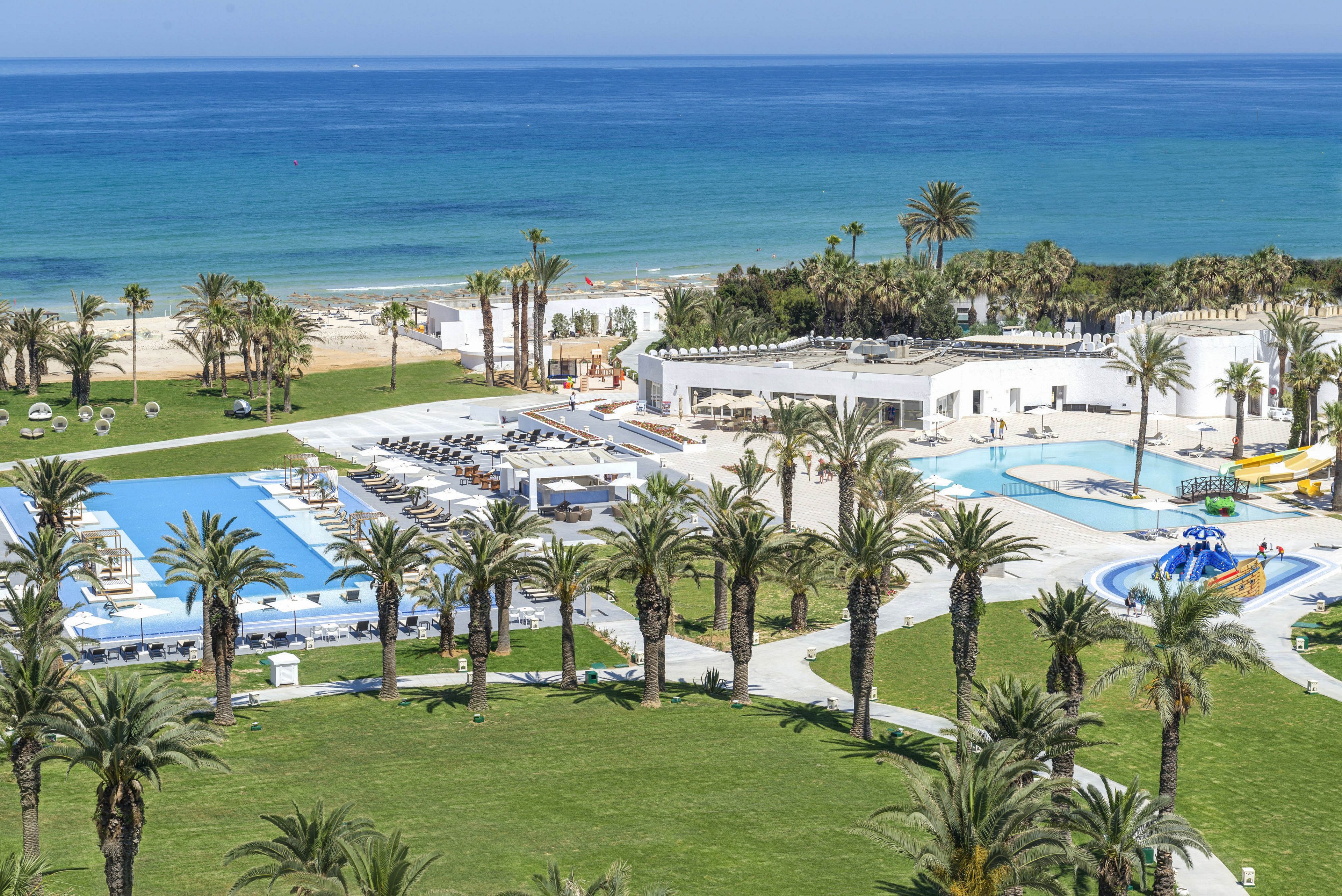 A coastal resort complex features multiple outdoor swimming pools surrounded by tall palm trees. White buildings and sun loungers line the pool decks, creating a bright and relaxing holiday atmosphere. Beyond the resort, a sandy beach leads to calm turquoise sea waters under a clear sky. The scene suggests a warm, sunny day ideal for vacation and leisure by the water.