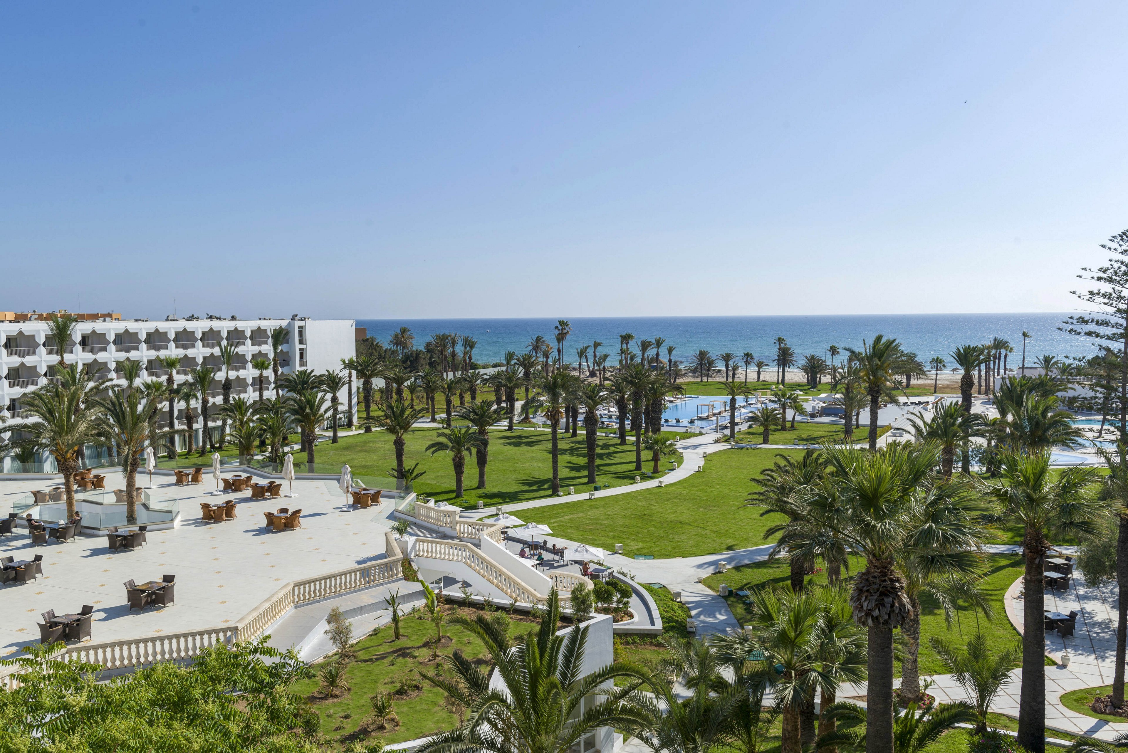 A large coastal resort overlooks manicured lawns and tall palm trees leading toward the sea. White multi-story hotel buildings frame expansive terraces furnished with outdoor seating areas. Curved pathways wind through green gardens toward swimming pools near the shoreline. The bright blue sky and calm ocean create a serene, sunny vacation atmosphere.