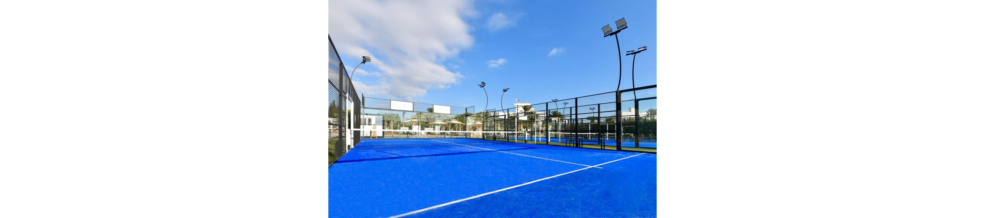 Image of an empty outdoor padel court with a bright blue playing surface. The court is enclosed by glass and metal fencing with tall light poles surrounding it. In the background, modern buildings, palm trees, and other sports facilities are visible. The scene is captured on a clear day with scattered clouds and strong natural light.