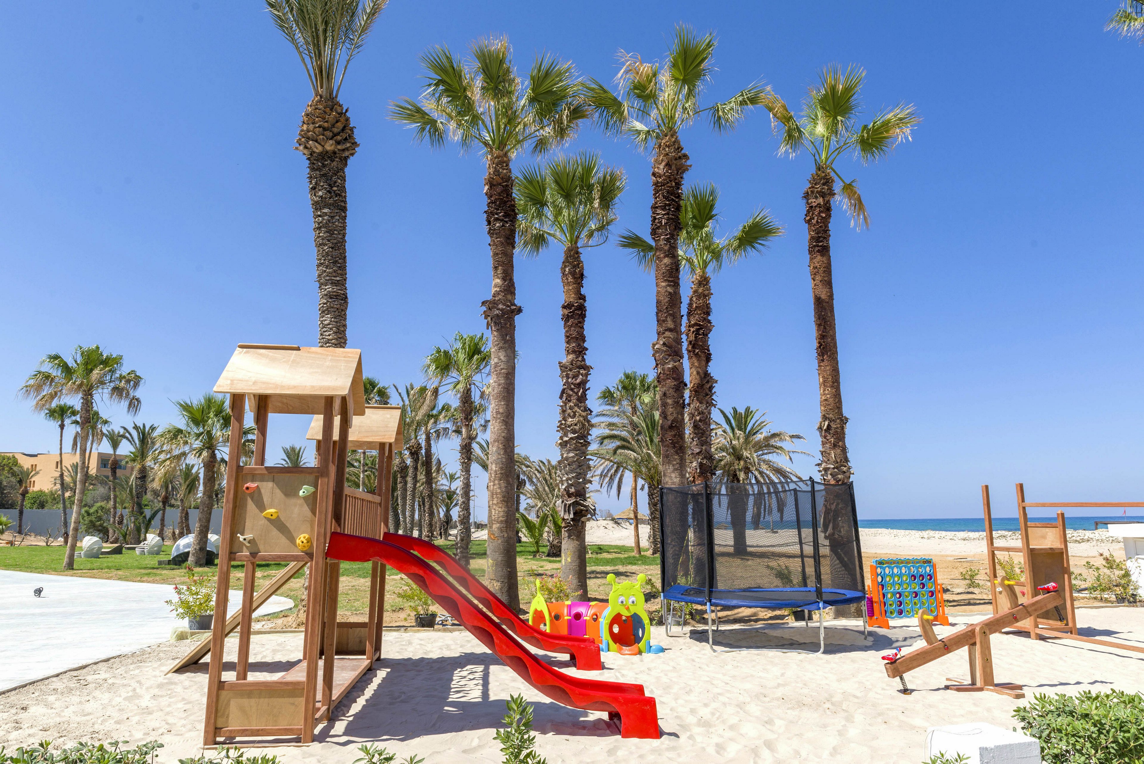 A colorful children's playground stands on sandy ground near a beach. The play area features a wooden climbing structure with a red slide, a trampoline, and several plastic ride-on toys. Tall palm trees rise in the background under a clear blue sky. The ocean and distant shoreline create a relaxed seaside resort atmosphere.