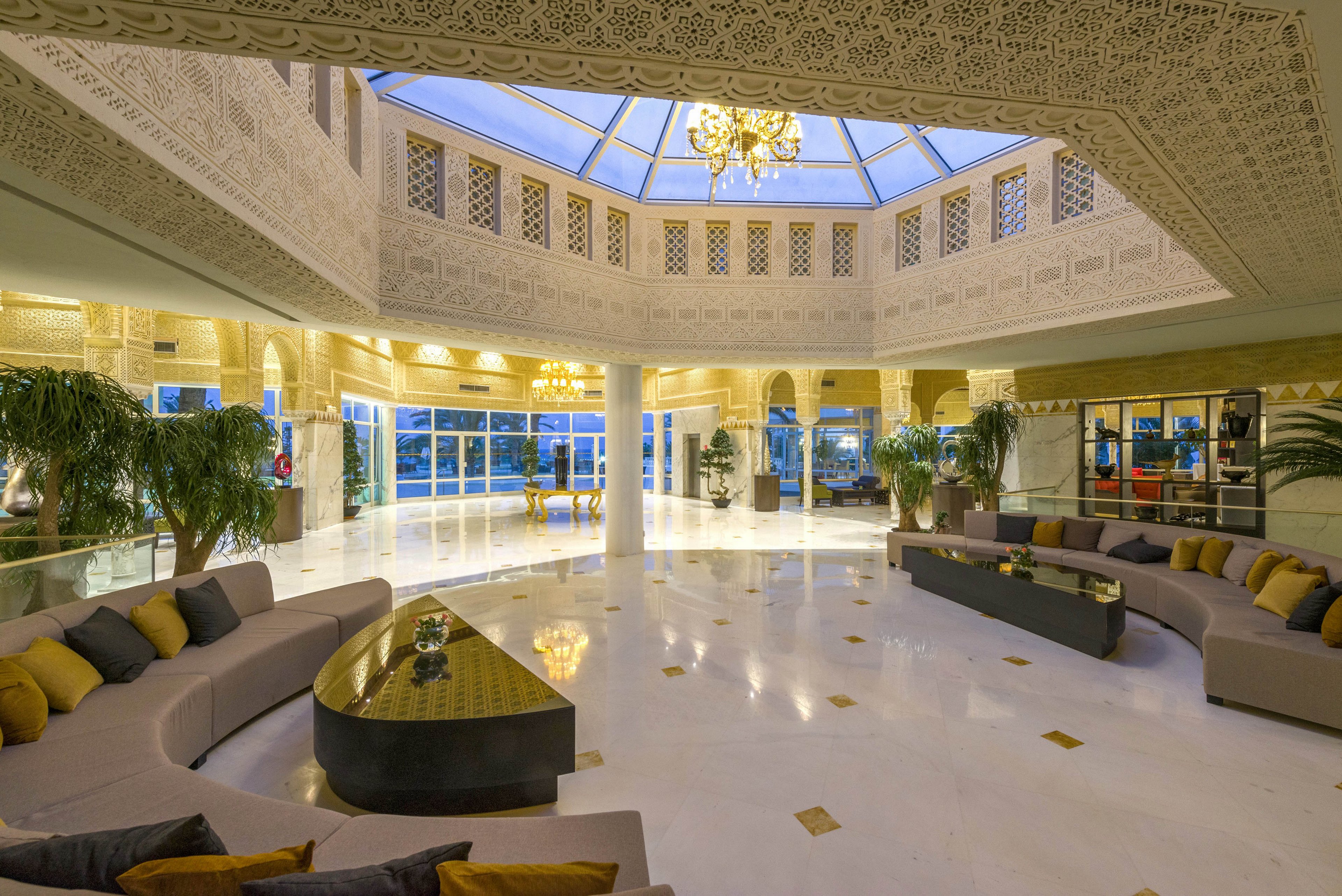 Spacious, elegant lobby interior featuring curved sectional sofas arranged around low coffee tables. The setting includes a large central skylight and intricately patterned walls and columns, suggesting a high-end hotel or resort. Warm lighting from chandeliers reflects off the polished marble floor, creating a sophisticated atmosphere. Large windows and glass doors open to an outdoor view, while indoor plants add a touch of greenery.