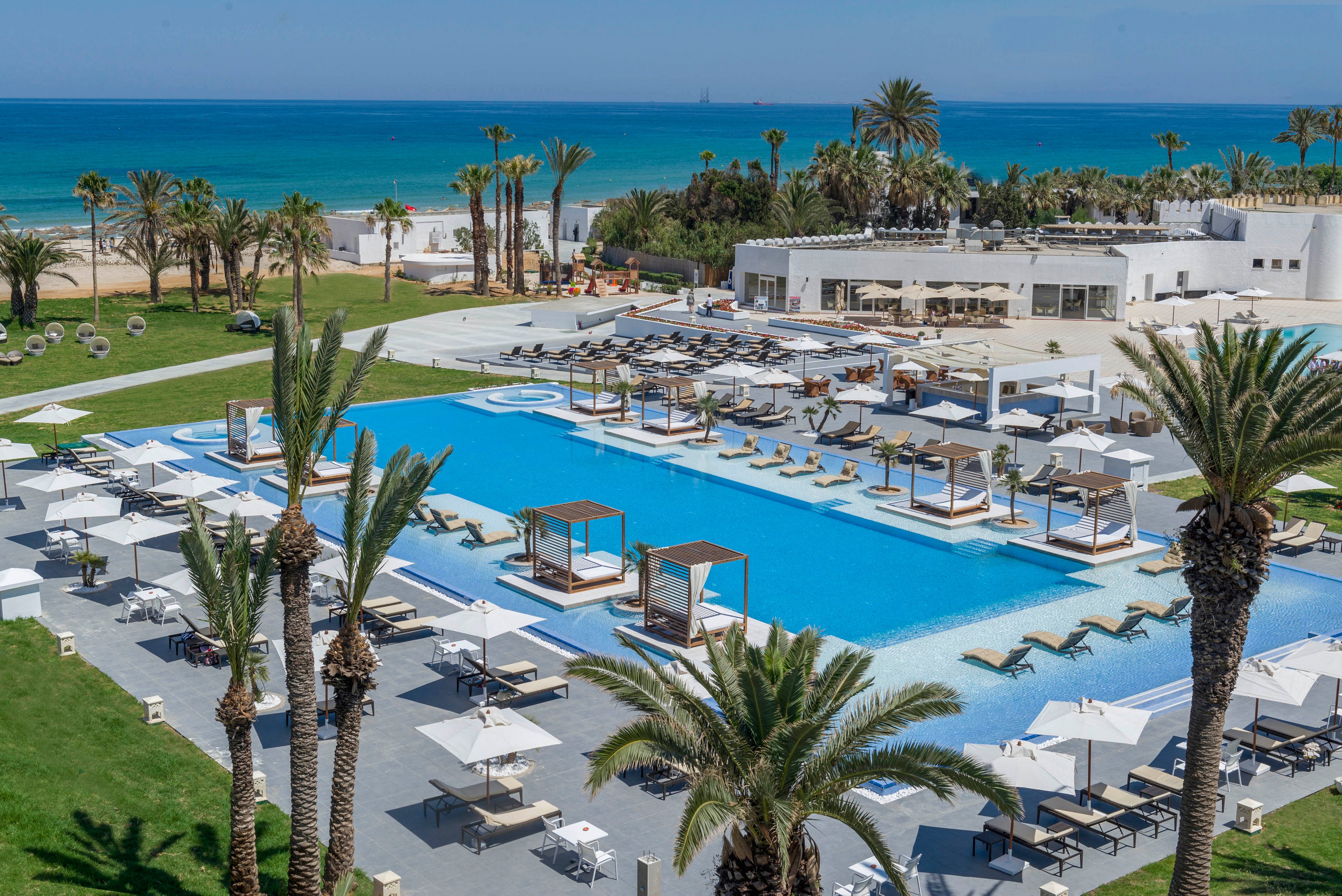 A modern beachfront resort features expansive infinity-style swimming pools surrounded by sun loungers and cabanas. White buildings and terraces overlook the pool area, with palm trees adding a tropical atmosphere. The turquoise sea stretches into the horizon under a clear blue sky. Guests relax on loungers and under umbrellas, creating a serene vacation setting.