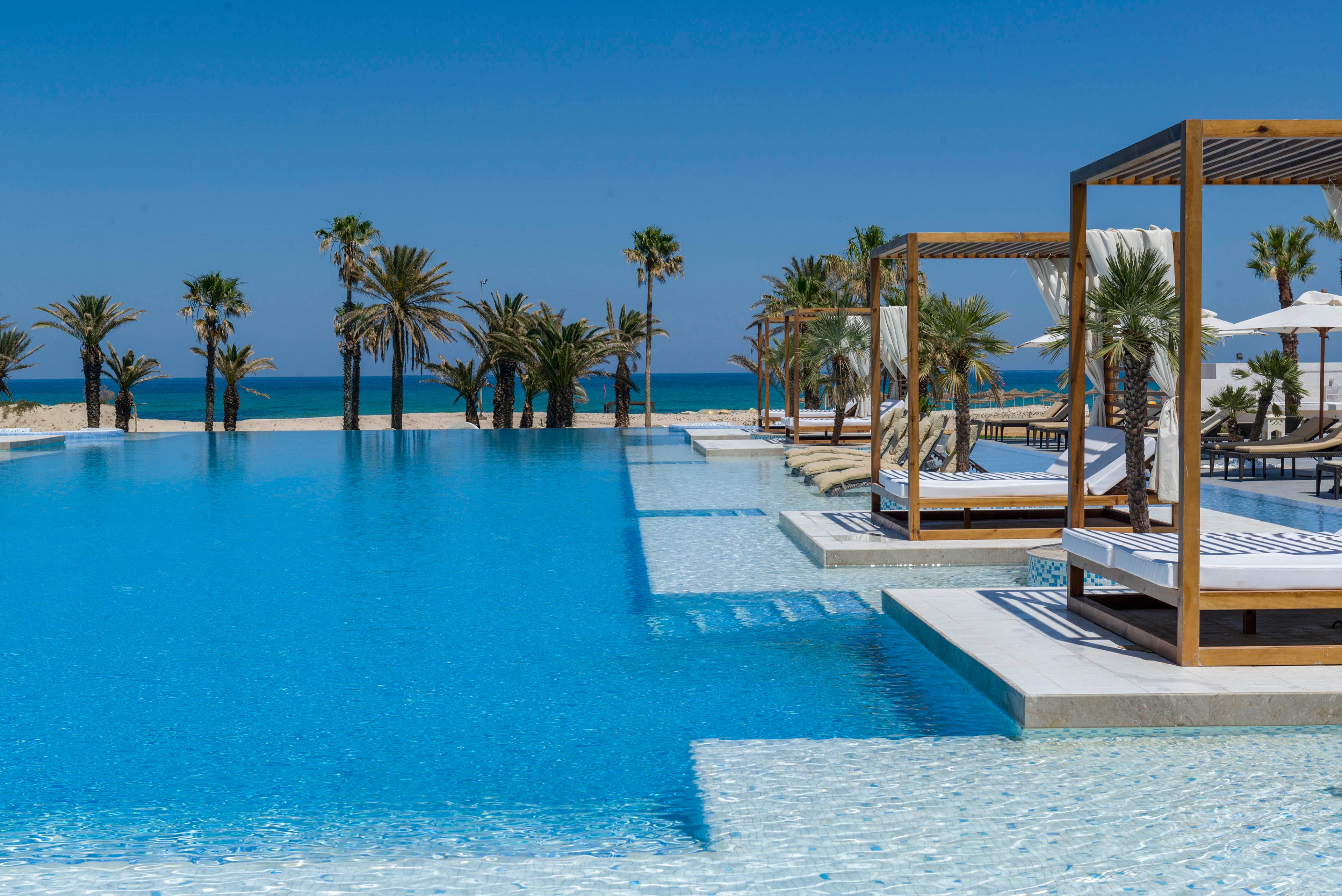 A large infinity pool stretches toward a sandy beach and calm blue sea. Wooden cabanas with white cushions line the poolside, offering shaded lounge areas. Tall palm trees frame the scene under a clear, cloudless sky. The tranquil setting evokes a high-end resort atmosphere ideal for relaxation and sunbathing.