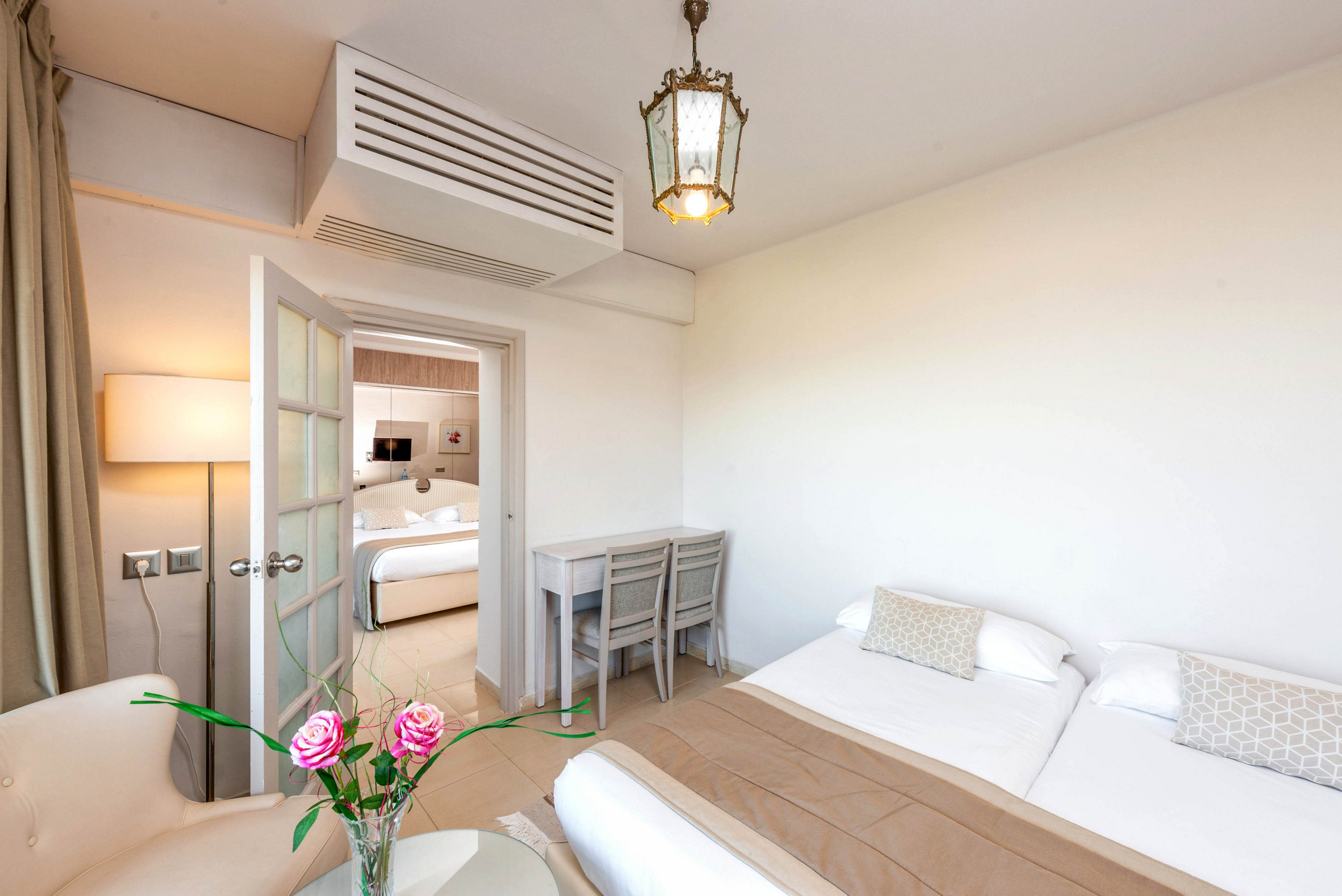 A bright hotel room features two neatly made twin beds with beige throws and patterned cushions. A small desk with two chairs sits against the wall near a glass-paneled door leading to an adjoining room. A vase with pink roses decorates a round table beside a white armchair, adding a touch of color. The space is lit by natural light and a hanging ceiling lamp, creating a clean and inviting atmosphere.