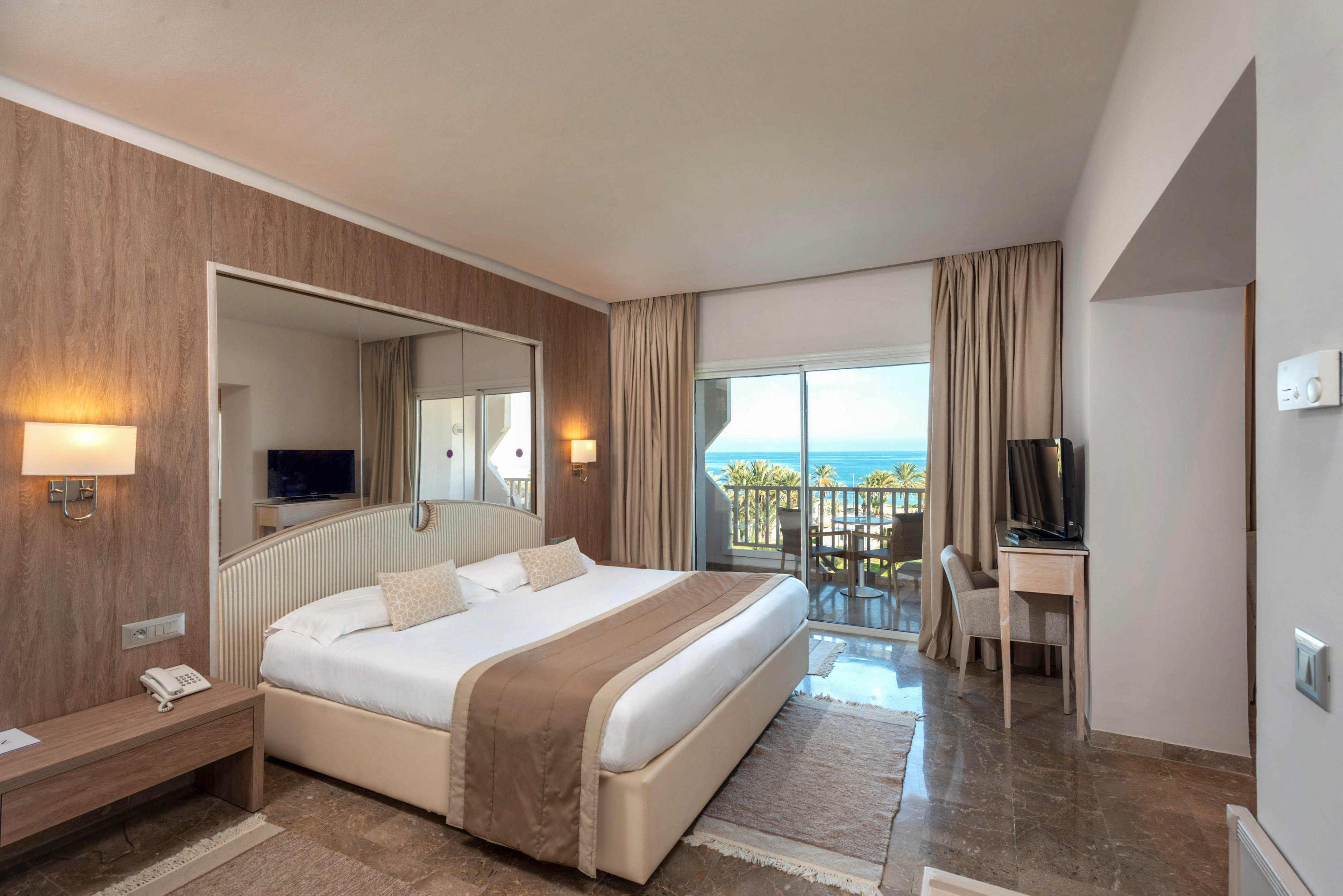 A spacious hotel room features a large double bed with neutral bedding and matching cushions. The interior includes wooden accents, bedside lamps, a wall mirror, and a small desk with a television. Floor-to-ceiling curtains frame sliding glass doors that open onto a balcony overlooking the sea and palm trees. The overall design is contemporary and minimal, creating a calm and relaxing atmosphere.