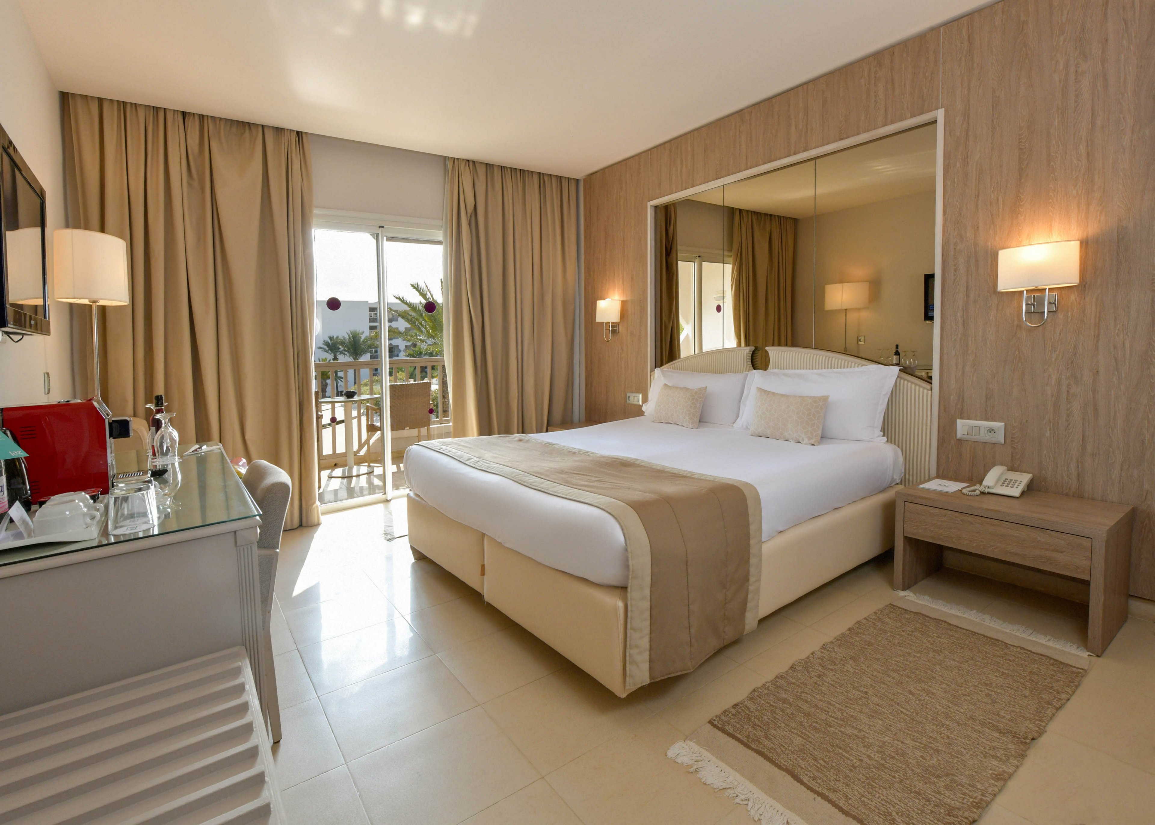 Contemporary hotel room featuring a large double bed with white linens and beige accents. The space includes a bedside table, wall-mounted lamps, and a desk with a coffee and tea station. Floor-to-ceiling curtains frame glass doors that open onto a balcony with outdoor seating and a partial sea view. Neutral tones and soft lighting create a calm, inviting atmosphere suitable for relaxation or business travel.
