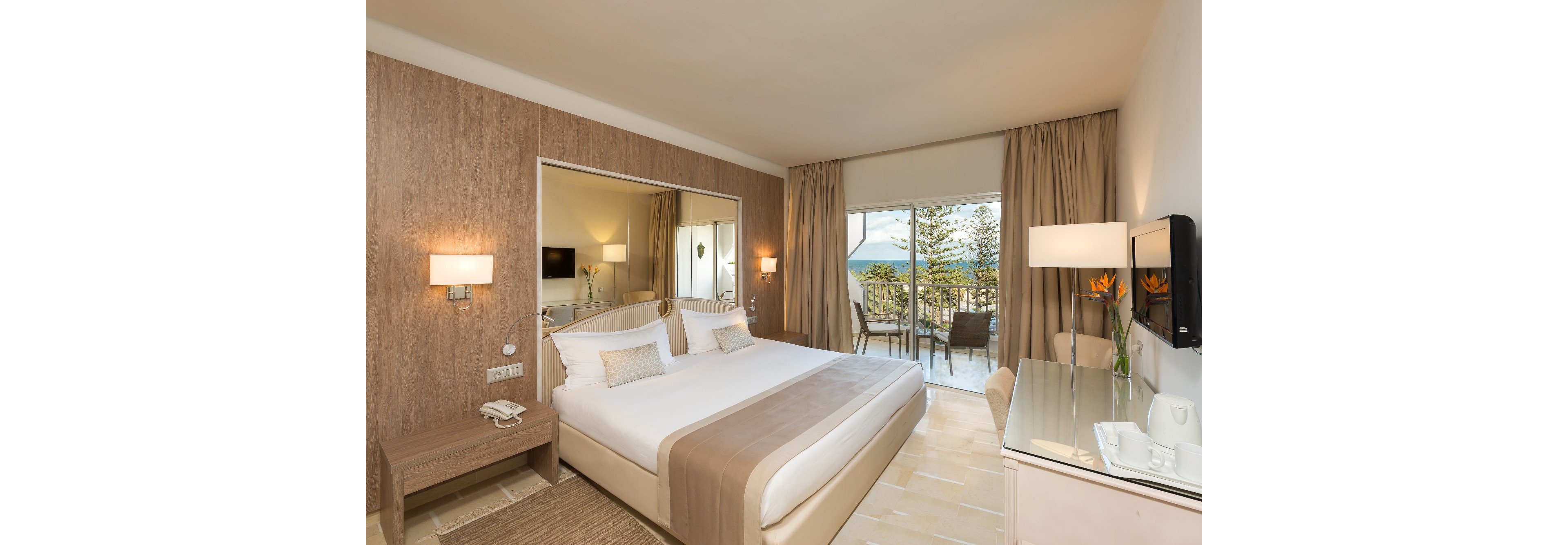 Modern hotel room featuring a large neatly made bed with neutral linens and wooden accents. A glass door opens to a balcony with chairs and a table overlooking trees and the sea. Soft beige tones, warm lighting, and simple decor create a calm, inviting atmosphere. A desk with a mirror, wall-mounted television, and fresh flowers complete the contemporary interior.
