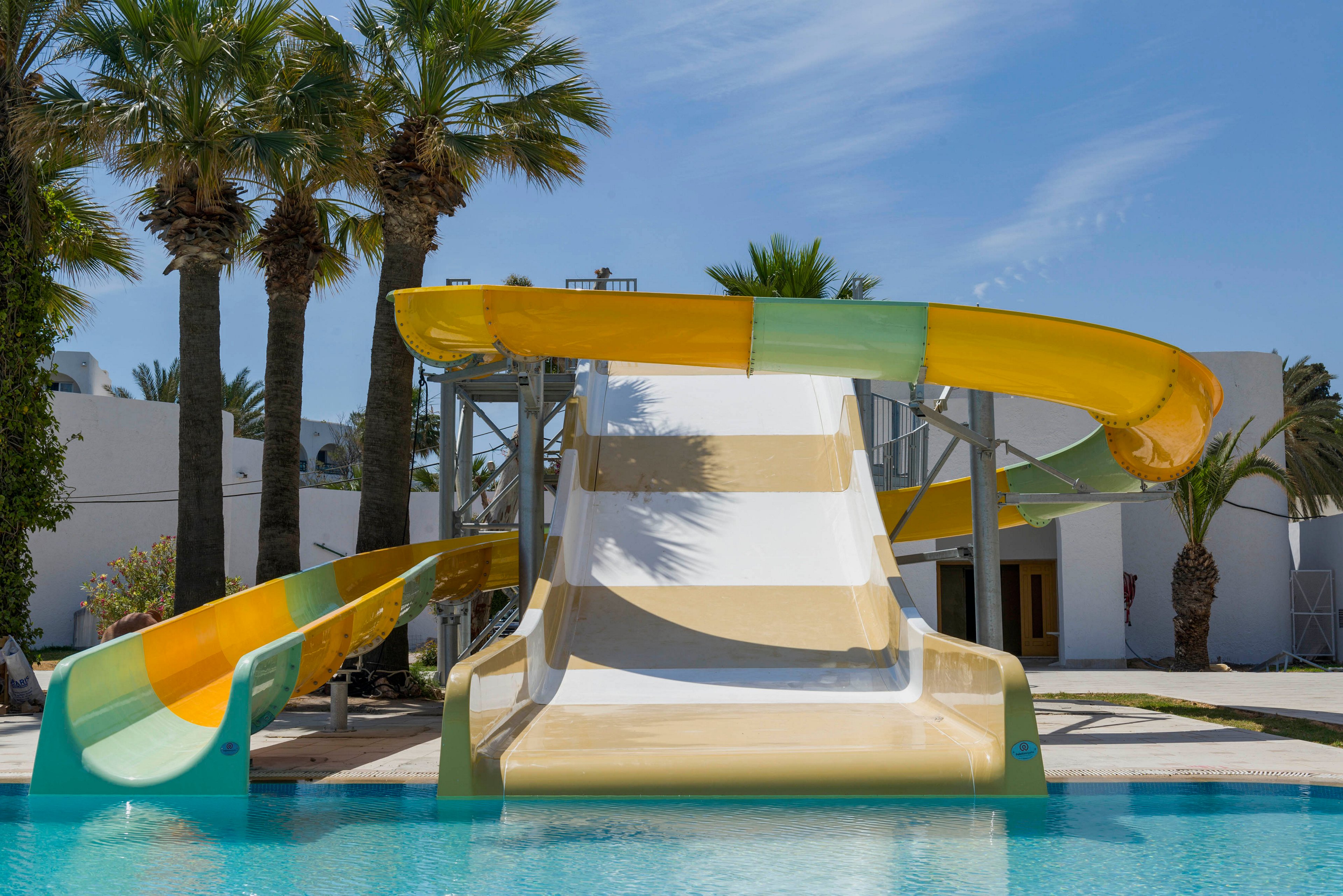 A large multi-lane water slide descends into a clear blue swimming pool at a sunny resort. The slide features wide white lanes bordered by yellow and green curved chutes. Tall palm trees and white modern buildings frame the background under a bright blue sky. The scene appears calm and inviting, with no people present.