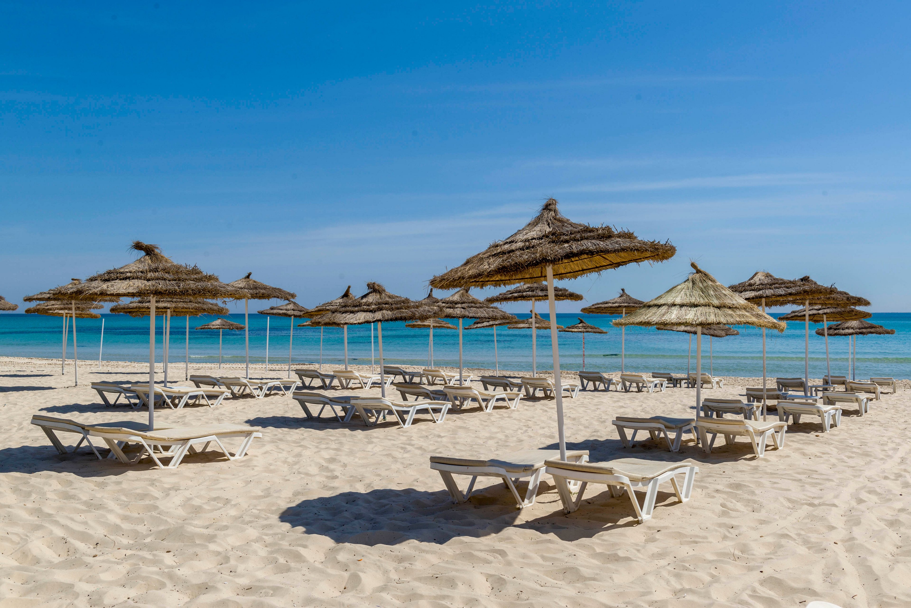 A sandy beach is lined with rows of white sun loungers under straw parasols. The scene is empty of people, creating a calm and peaceful atmosphere. Turquoise water gently meets the shore under a clear blue sky. The image evokes a relaxing seaside holiday destination.