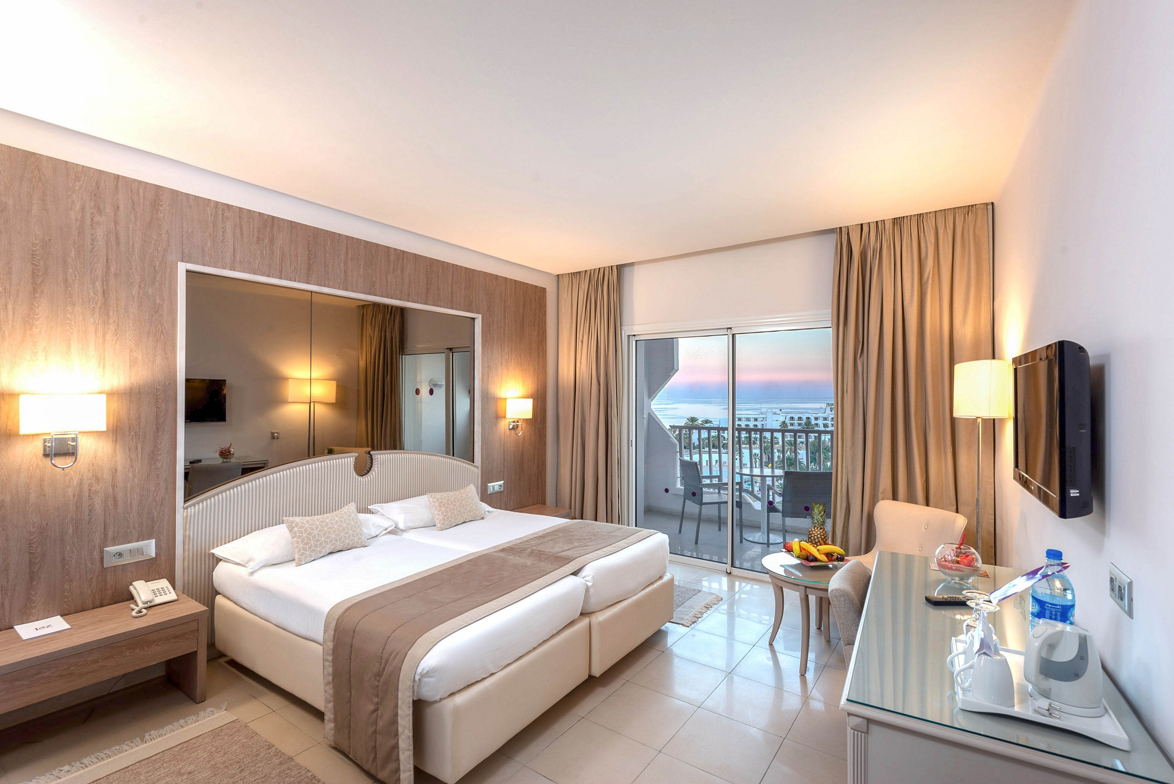 Bright contemporary hotel room featuring a large double bed with neutral bedding and cushions. A wide mirror behind the bed and soft wall lamps create a spacious, inviting atmosphere. A desk with a television, kettle, and bottled water sits opposite a small seating area with a round table holding fruit and drinks. Sliding glass doors open to a balcony overlooking the sea at sunset, adding a relaxing vacation feel.