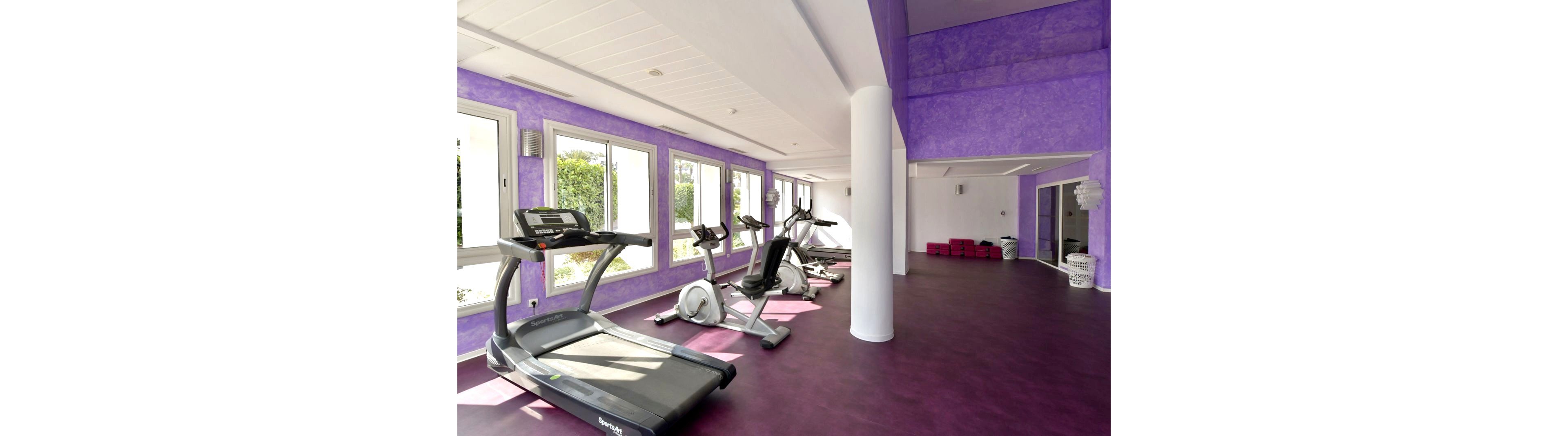 A contemporary indoor gym features several cardio machines, including treadmills and stationary bikes, arranged along large windows. Natural light floods the space, highlighting the purple walls and matching floor. White columns and ceiling panels create a clean, open atmosphere. In the background, red exercise blocks and fitness accessories add contrast to the colorful room.