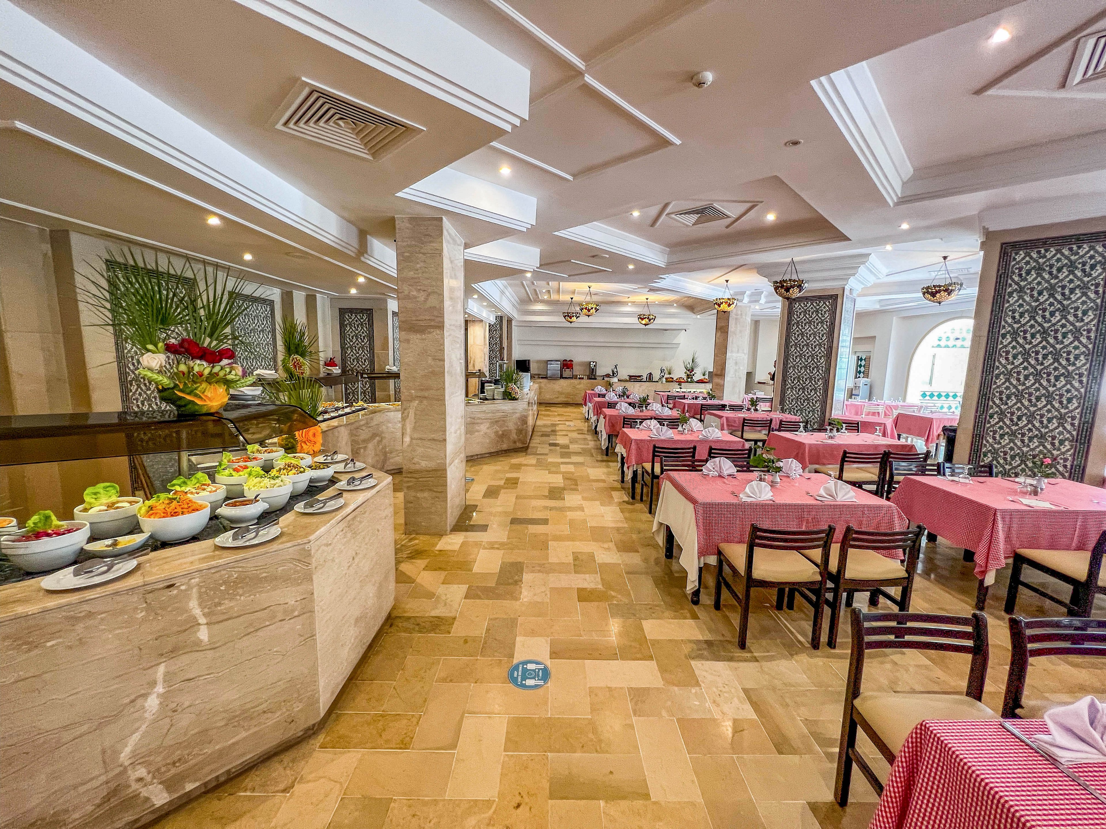 Spacious hotel restaurant interior features a buffet counter with assorted fresh fruits and salads in white bowls. Rows of dining tables covered in pink and white checkered tablecloths extend through the room, paired with dark wooden chairs. Decorative ceiling details, hanging chandeliers, and patterned wall panels add an elegant touch. The tiled floor and natural light from arched windows create a warm, inviting atmosphere for guests.