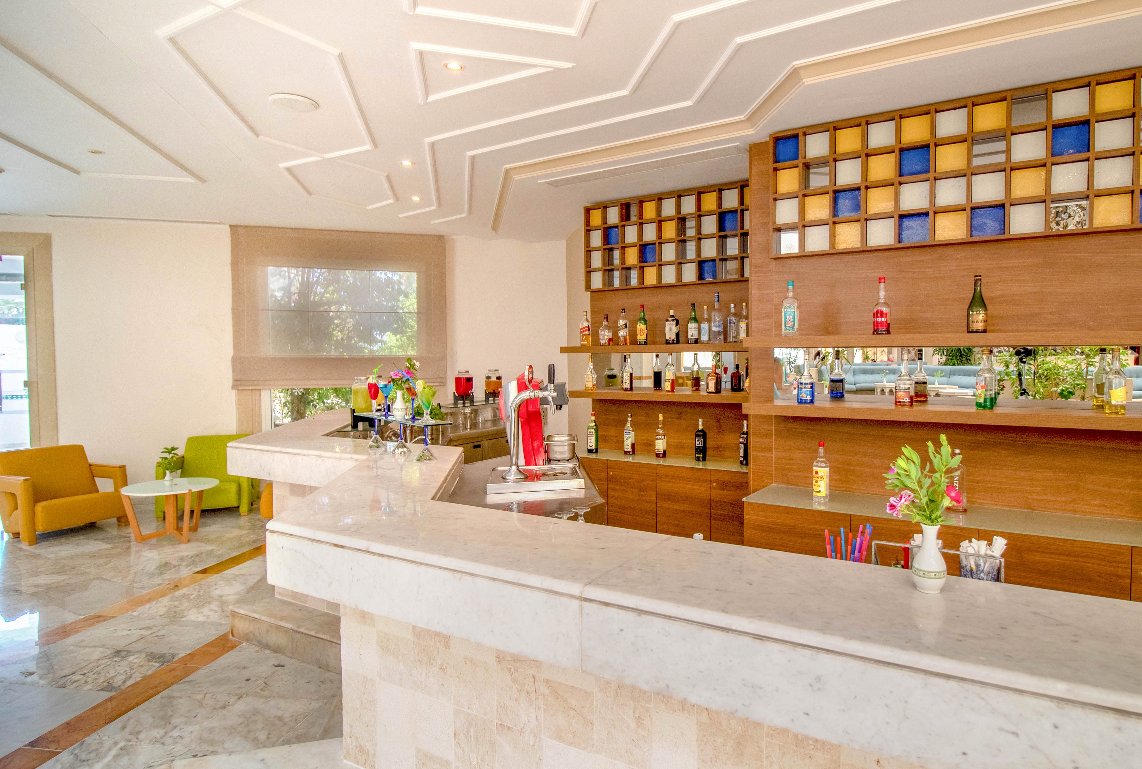 A contemporary hotel bar features a long marble counter with various bottles and glassware neatly arranged. The back wall showcases wooden shelving with colorful square accents and a selection of spirits. Fresh flowers and bar tools add decorative touches to the clean, well-lit space. Comfortable seating and large windows create an inviting atmosphere for guests.