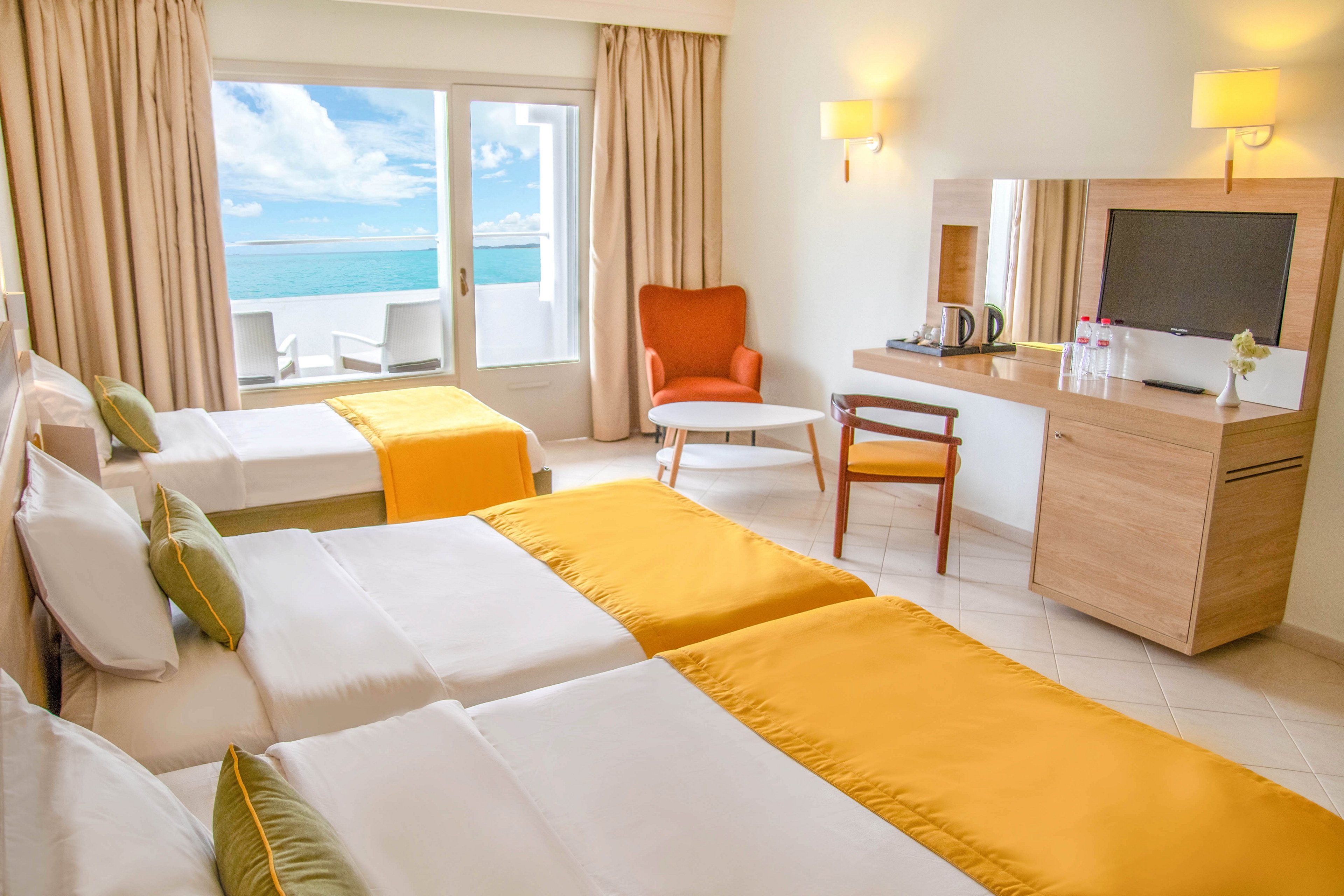 A modern hotel room features two twin beds with white linens and vibrant yellow bed runners. The space is decorated in light neutral tones with green accent pillows, a wooden desk, and a cozy orange armchair. Large glass doors open to a balcony with a view of the ocean and blue sky. Soft natural light and simple contemporary furnishings create a clean, inviting atmosphere.