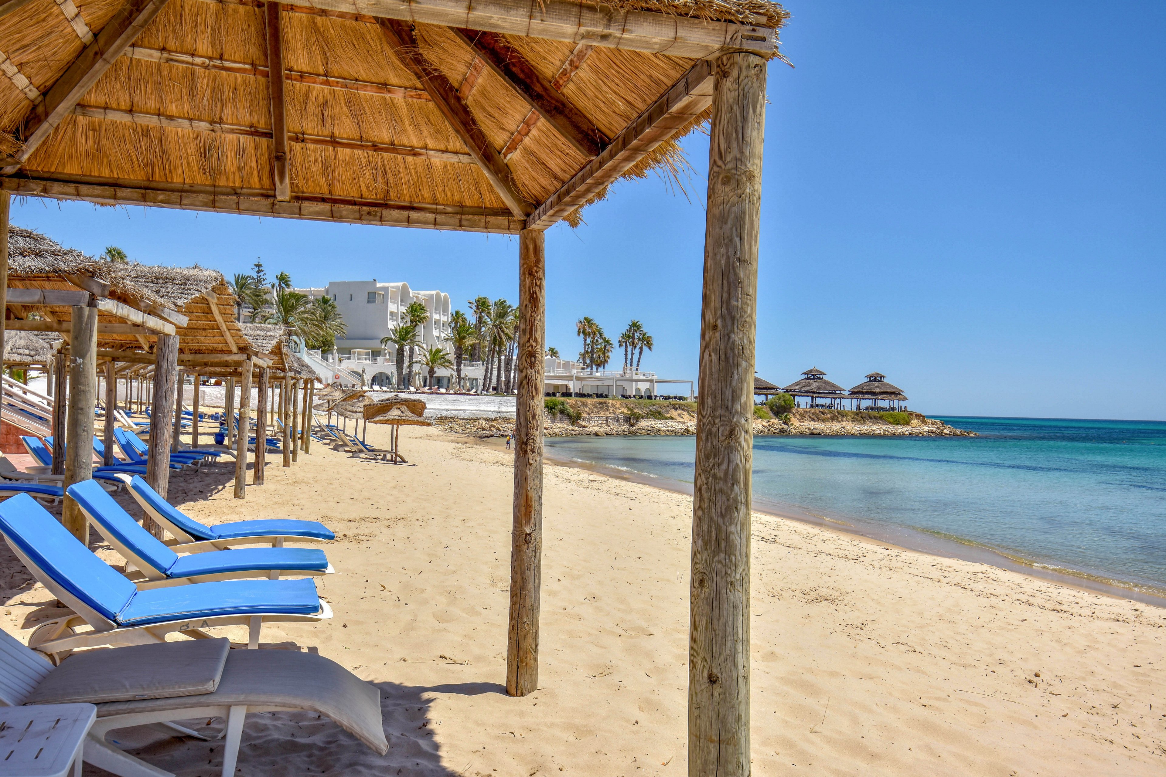 A row of blue sun loungers sits under thatched shade structures on a sandy beach beside clear blue water. In the background, white resort buildings and palm trees line the shoreline, suggesting a tranquil holiday destination. The sky is bright and cloudless, emphasizing the warm, sunny weather. The scene conveys relaxation and leisure at a coastal resort.