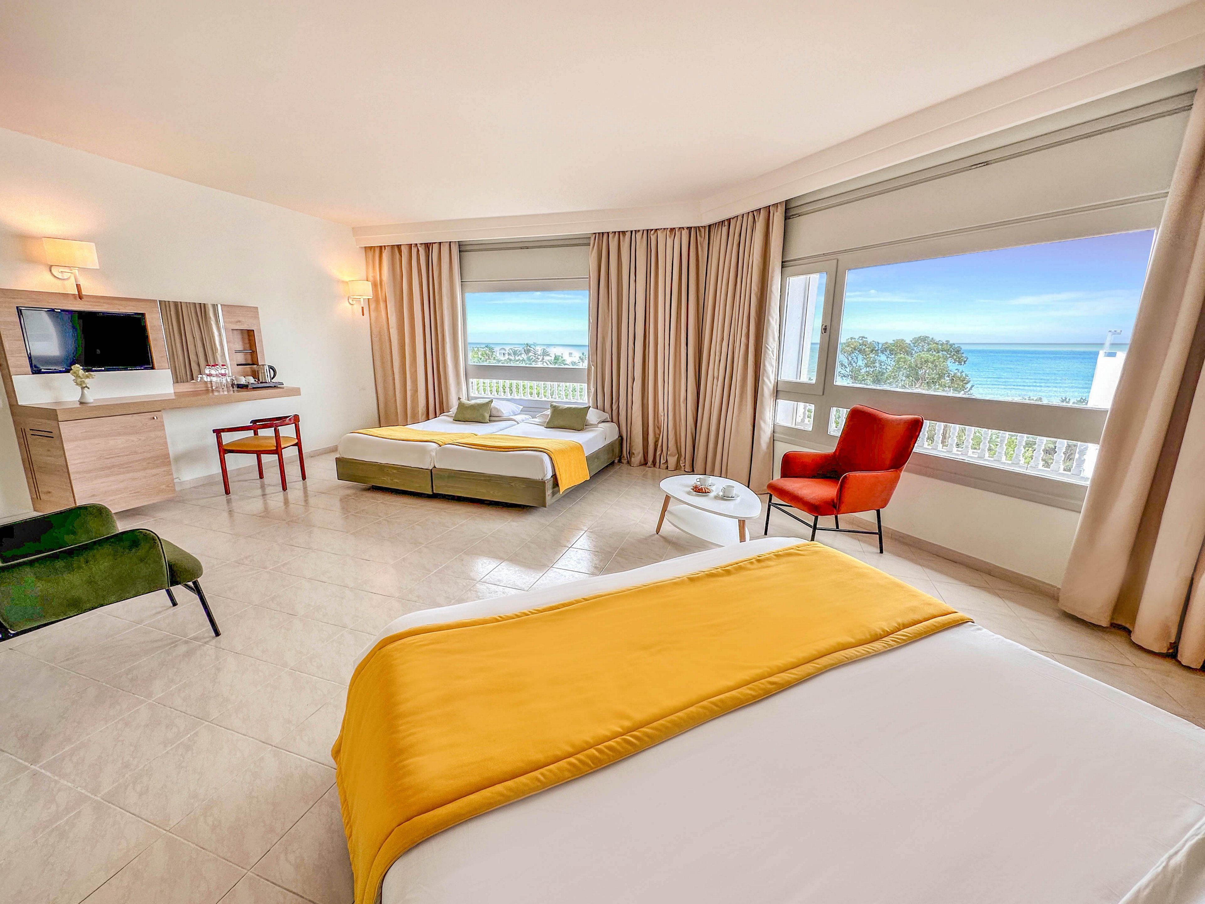Spacious hotel room features a large bed in the foreground and two single beds near wide windows. Neutral walls and tiled floors are accented by yellow bed throws, beige curtains, and colorful seating including a red armchair and green bench. A desk area with a wall-mounted television and kettle sits to the left, adding functional comfort. Large windows reveal a scenic ocean view and blue sky, filling the room with natural light.