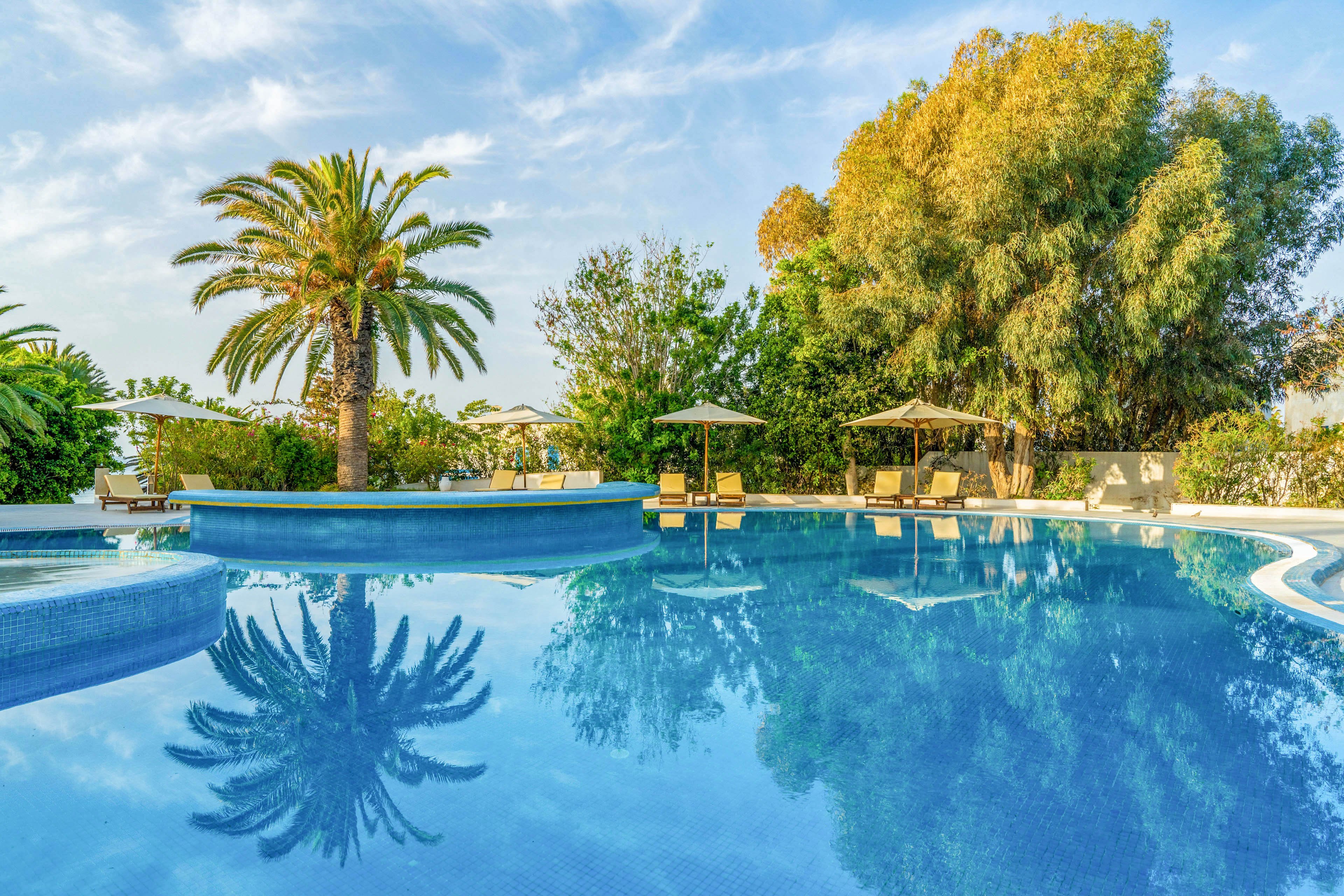 A clear blue swimming pool reflects surrounding palm trees and lush greenery under a bright sky. Sun loungers and parasols are arranged around the poolside, suggesting a relaxing vacation setting. The calm water and warm light create a tranquil, inviting atmosphere. The scene evokes a Mediterranean or tropical resort environment focused on leisure and relaxation.