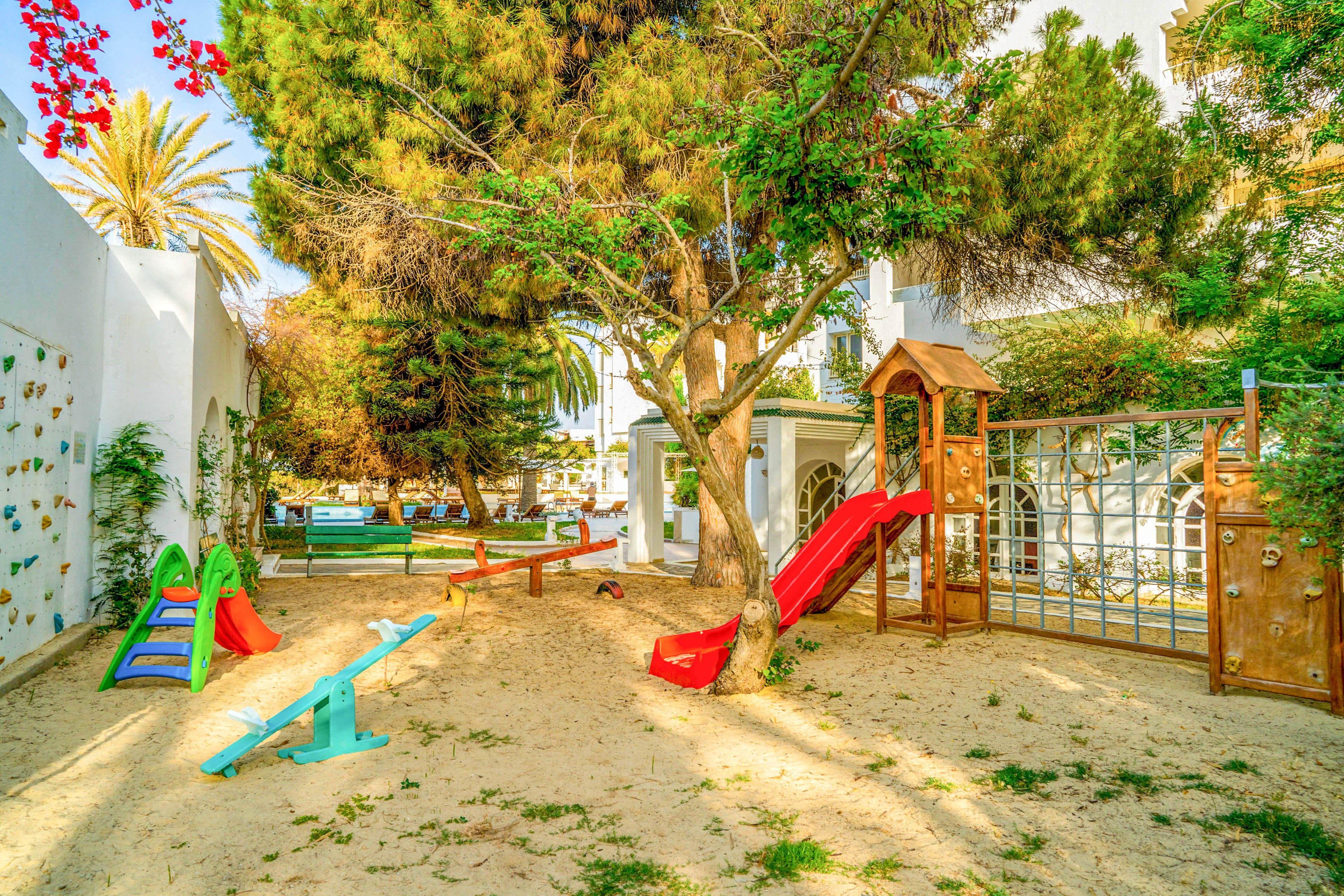An outdoor playground is set in a sandy yard surrounded by lush green trees and white walls. Colorful play equipment, including slides, a seesaw, and climbing structures, creates a fun and inviting space for children. Sunlight filters through the trees, casting warm shadows across the sand and play area. The scene suggests a relaxed, family-friendly environment in a residential or holiday setting.