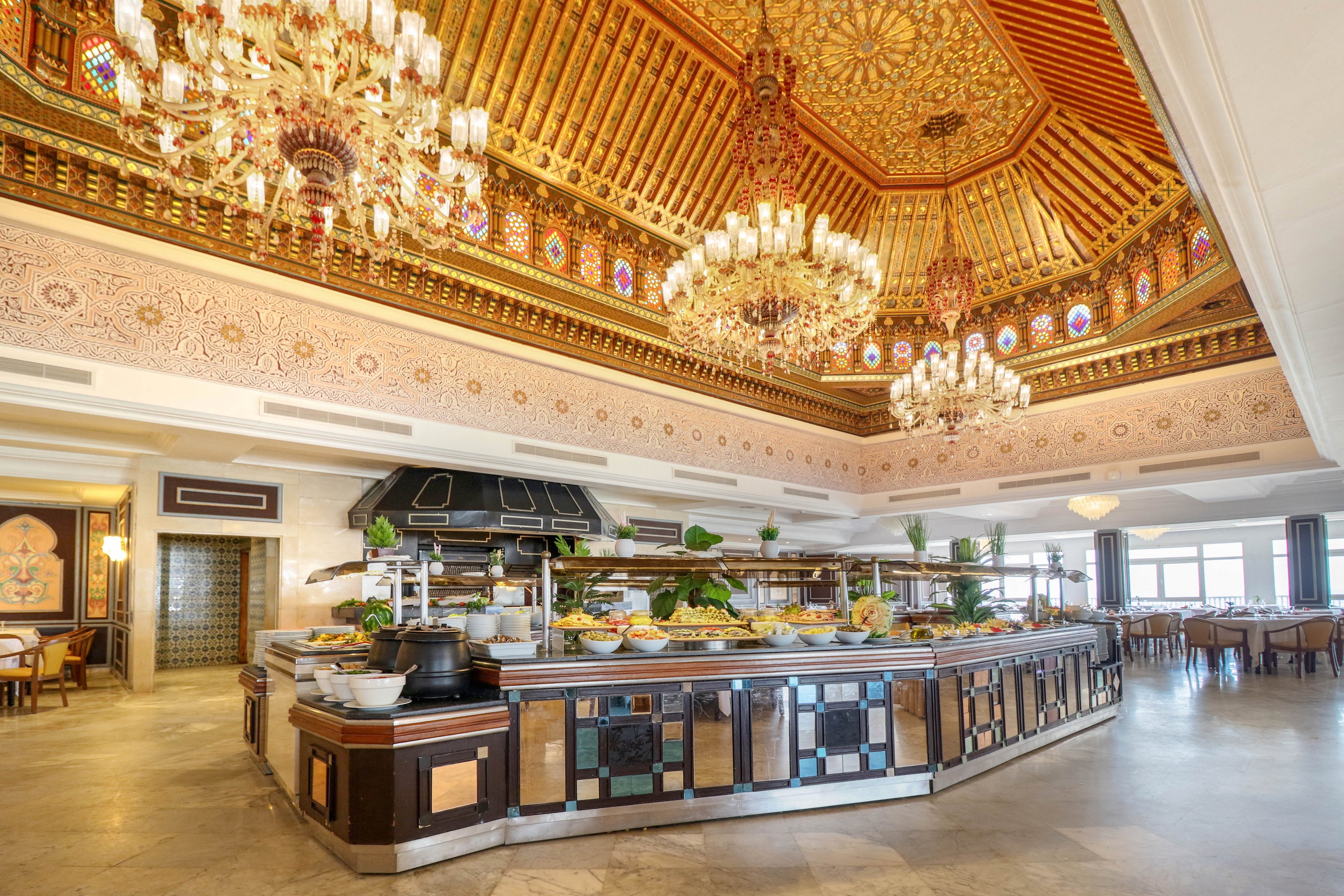 An opulent buffet station stands in the center of a grand restaurant interior. The ceiling features intricate golden patterns, stained-glass details, and multiple crystal chandeliers. Various dishes and fresh greens are displayed along the counter, ready for guests. Elegant seating areas and large windows create a bright, spacious dining atmosphere.