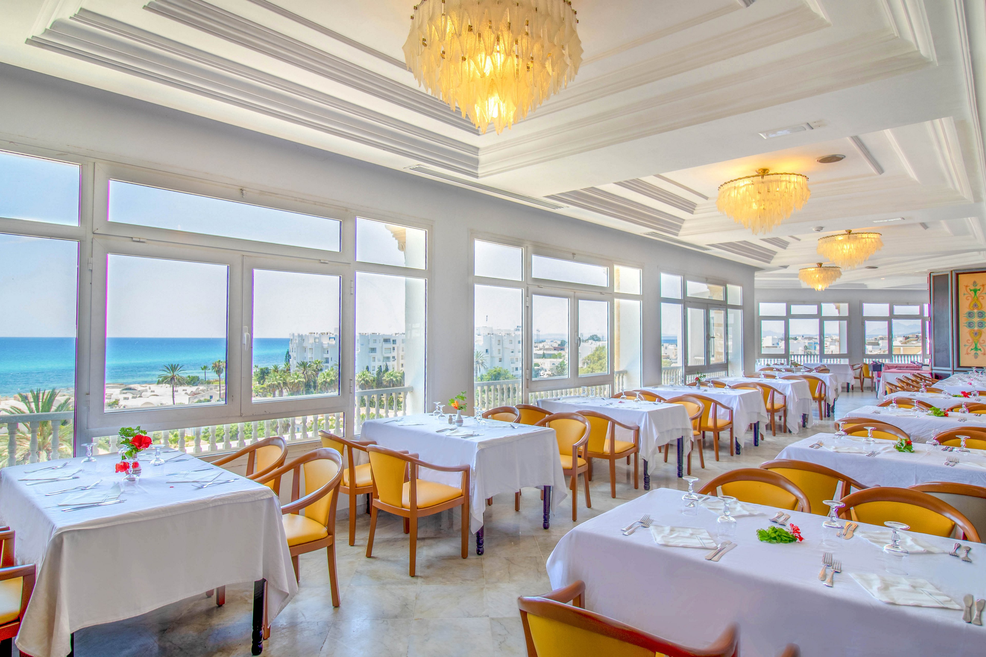 A spacious, sunlit restaurant features neatly arranged tables covered with white tablecloths and simple place settings. Large windows line the room, offering expansive views of the blue ocean and coastal buildings outside. Warm yellow chairs and crystal chandeliers add a touch of elegance to the airy interior. Small floral centerpieces provide colorful accents against the predominantly white and light-toned decor.