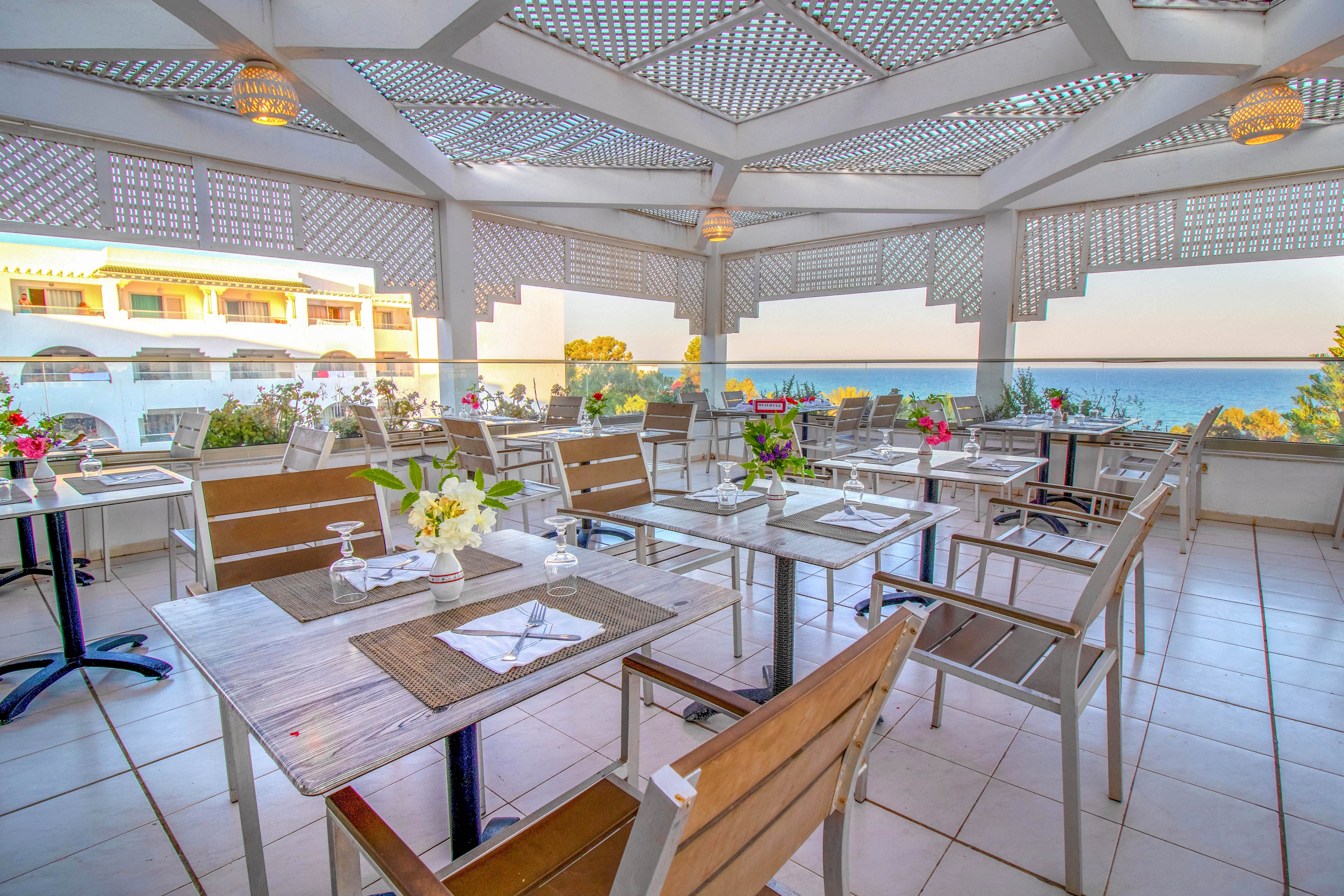 An open-air restaurant terrace features neatly arranged tables with wooden chairs and placemats. Fresh flowers and glassware decorate each table, creating an inviting dining atmosphere. The space is covered by a patterned pergola roof that filters the sunlight. In the background, a white building and lush greenery frame a clear view of the sea at sunset.