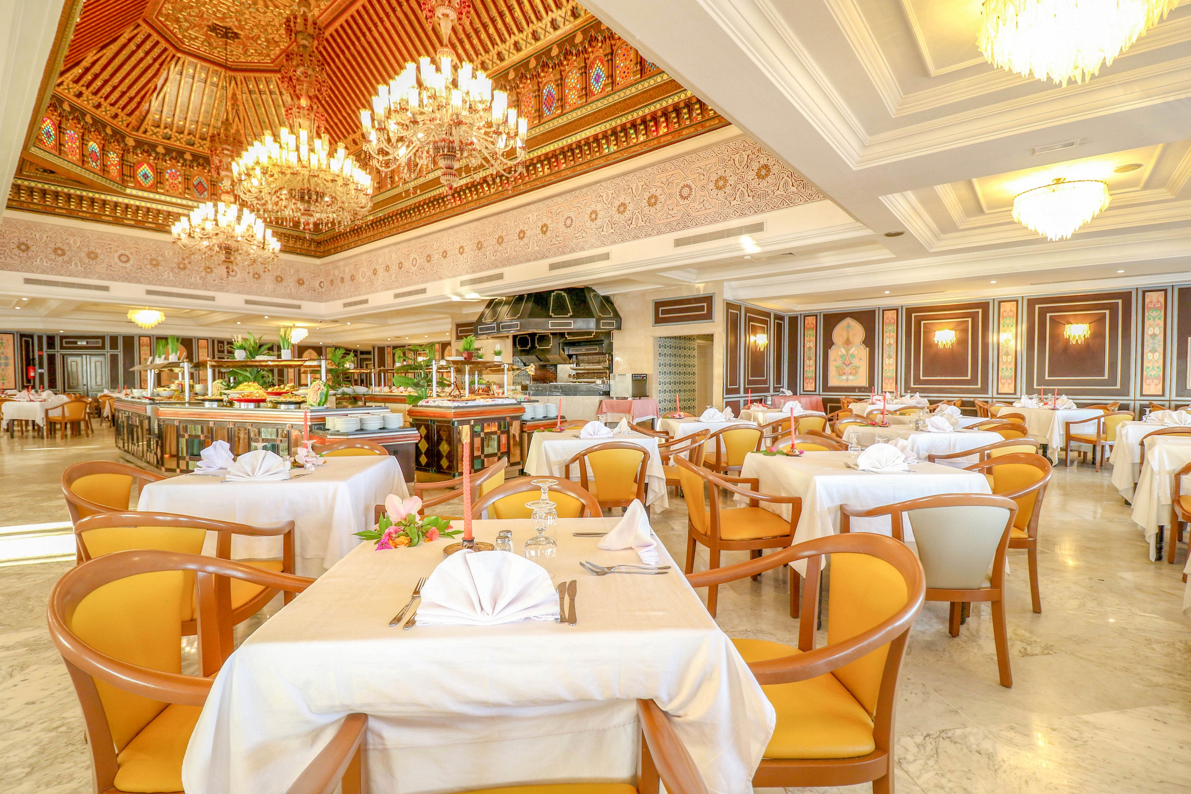 Spacious upscale restaurant interior with neatly arranged tables and chairs. White tablecloths and folded napkins create a formal dining atmosphere, complemented by warm wooden seating. Large ornate chandeliers hang from a richly decorated ceiling, casting a golden glow across the room. Decorative wall panels and framed artwork enhance the luxurious, classic style of the space.