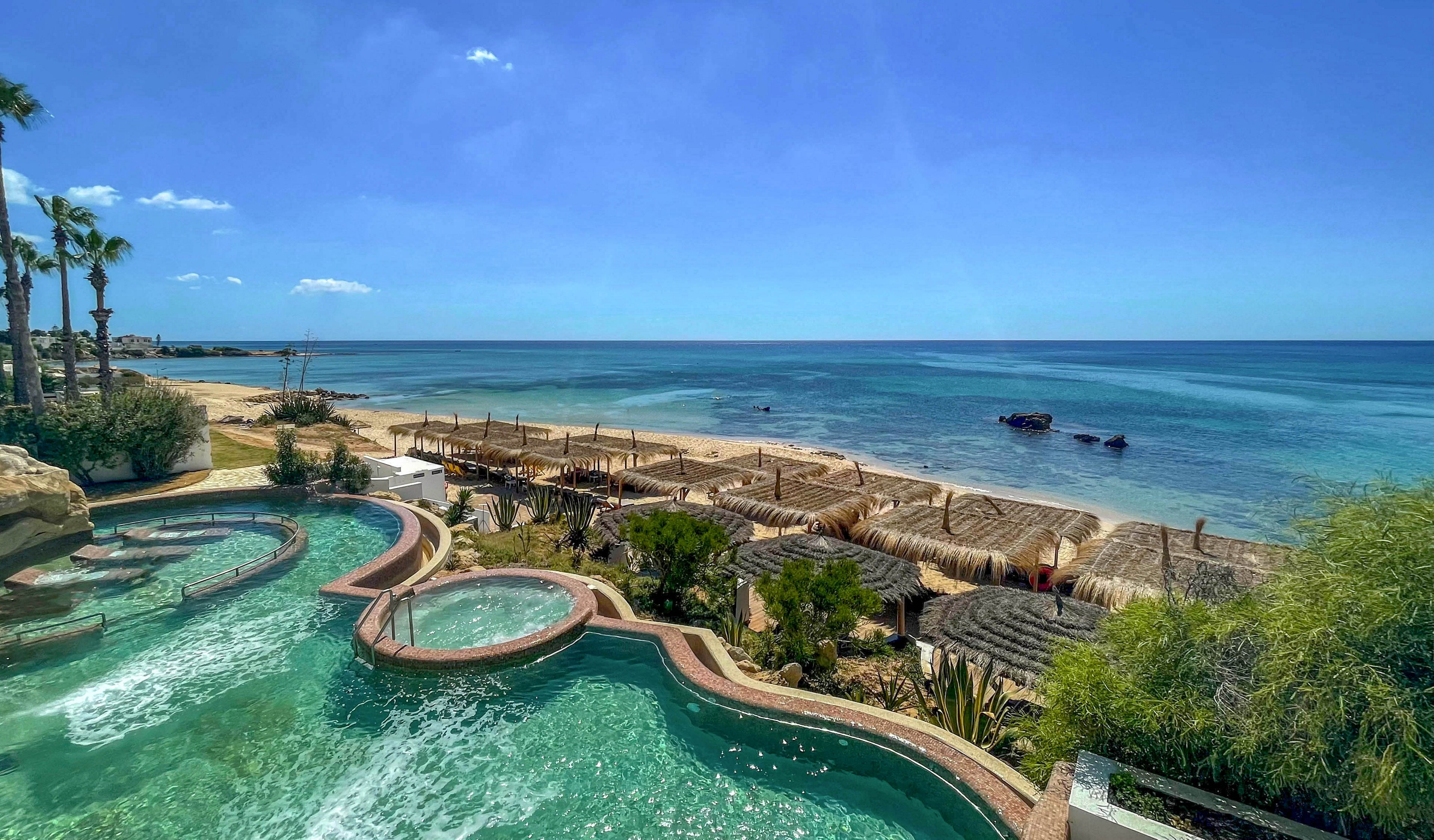 A turquoise resort pool with curved edges overlooks a sandy beach and calm blue ocean. Thatched cabanas and shaded lounge areas line the shoreline, creating a tropical vacation atmosphere. Palm trees and lush greenery frame the scene under a clear, sunny sky. The overall setting suggests a relaxing coastal getaway at an upscale seaside resort.
