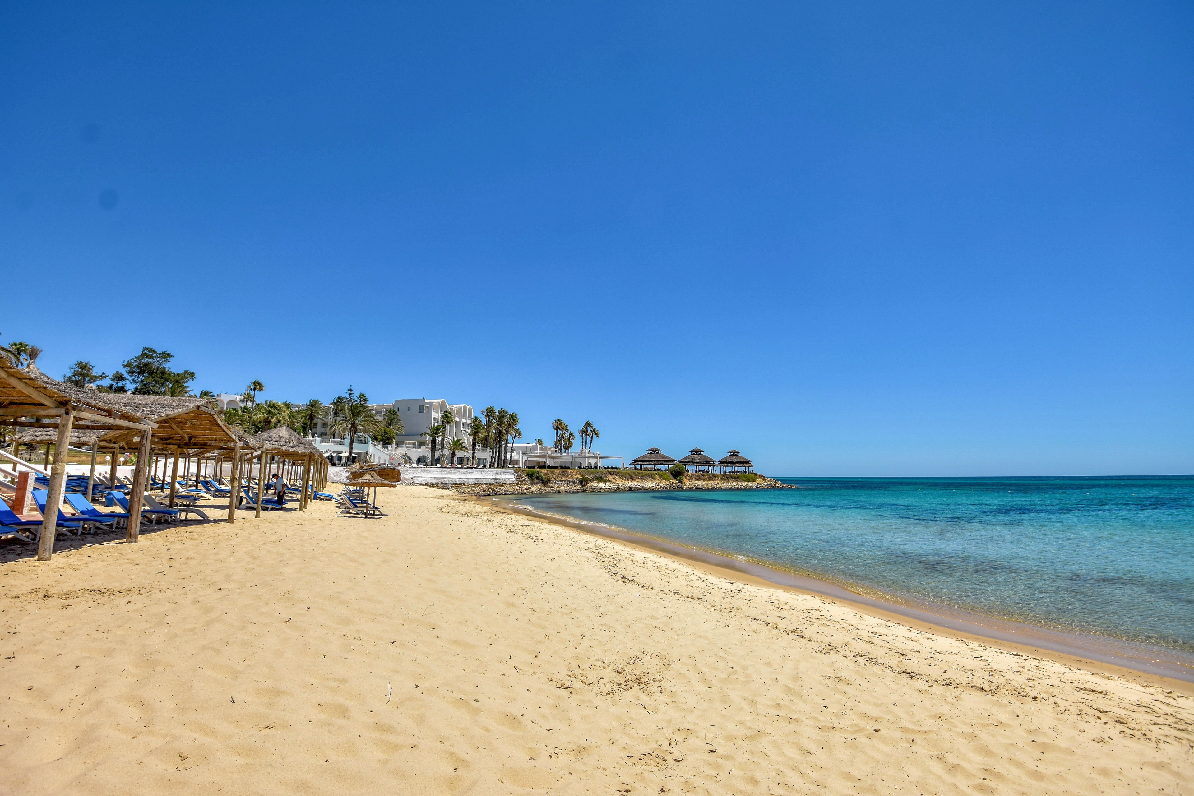 A wide sandy beach stretches along a calm, clear blue sea under a bright cloudless sky. Straw umbrellas and lounge chairs line the shoreline, creating a relaxed vacation setting. In the distance, palm trees and low white buildings suggest a coastal resort area. The scene feels peaceful and inviting, ideal for sunbathing and seaside holidays.