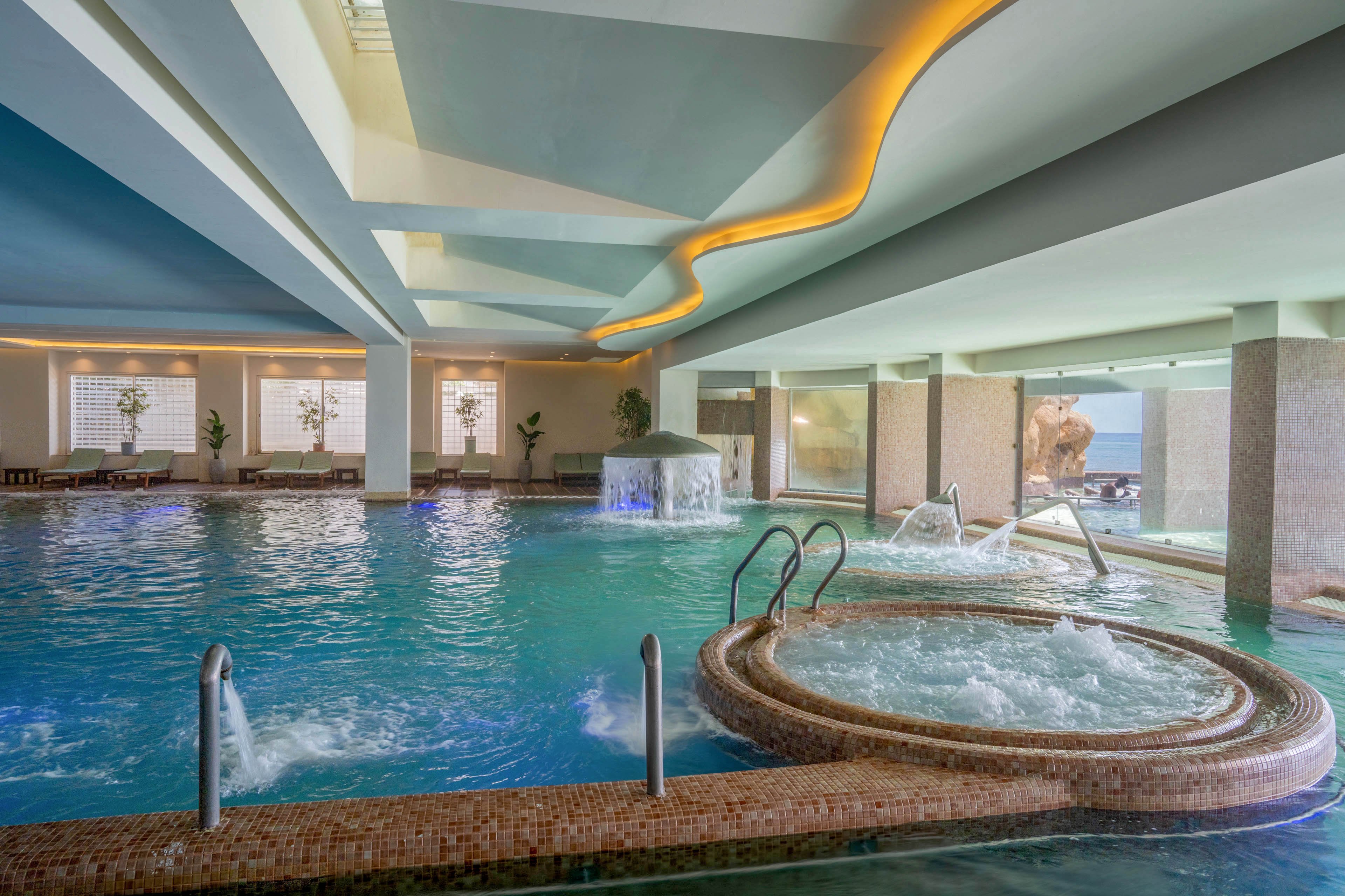 Spacious indoor spa pool area featuring multiple hydrotherapy jets and a circular whirlpool section. Water cascades from a fountain-like structure into the main pool, creating a relaxing atmosphere. Lounge chairs line the far side of the room, with large windows letting in natural light and a view toward the sea. Soft ceiling lighting and modern architectural details enhance the tranquil, resort-style setting.