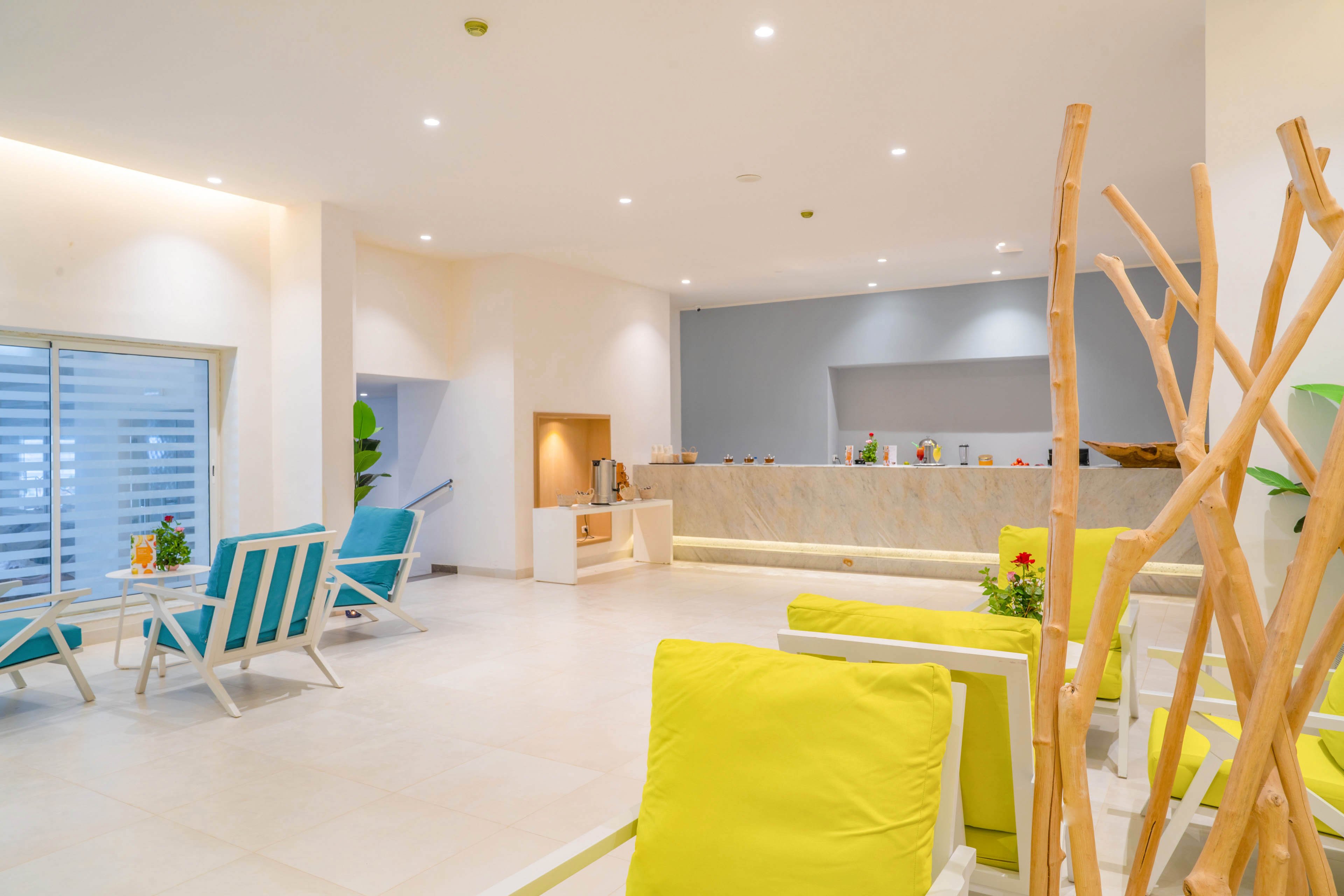 A contemporary hotel lobby features colorful lounge chairs and a sleek reception desk. The space is open and airy, with white walls, light flooring, and recessed ceiling lights. Turquoise and yellow cushions add a vibrant contrast to the neutral surroundings. Wooden decorative elements and potted plants provide a warm, welcoming atmosphere.