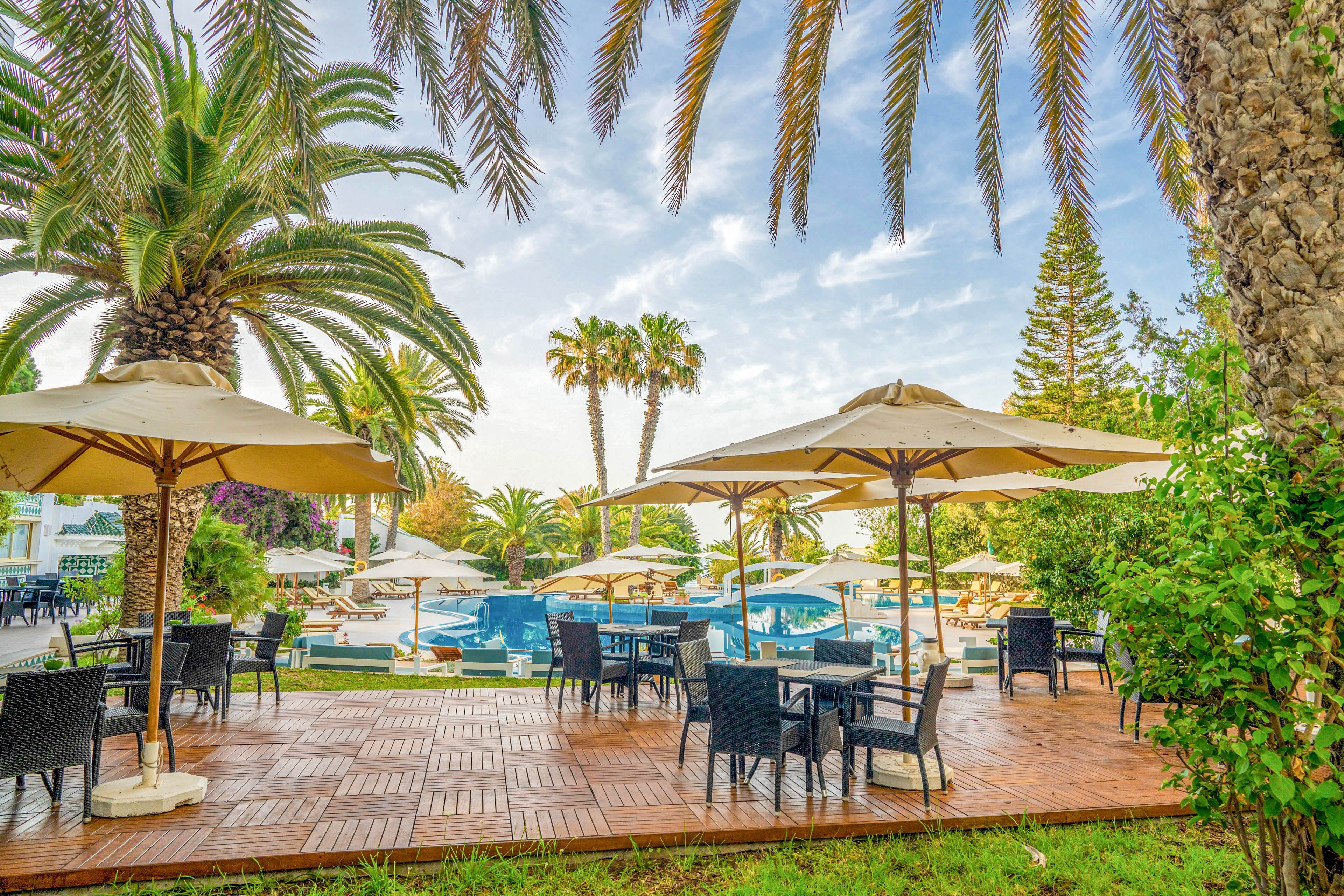 An inviting outdoor terrace at a resort features tables and chairs arranged beneath large beige umbrellas. Beyond the wooden deck, a swimming pool area is surrounded by sun loungers and more umbrellas. Tall palm trees and lush greenery frame the scene, creating a tropical holiday atmosphere. The bright daylight and soft blue sky suggest a warm, relaxing vacation setting.