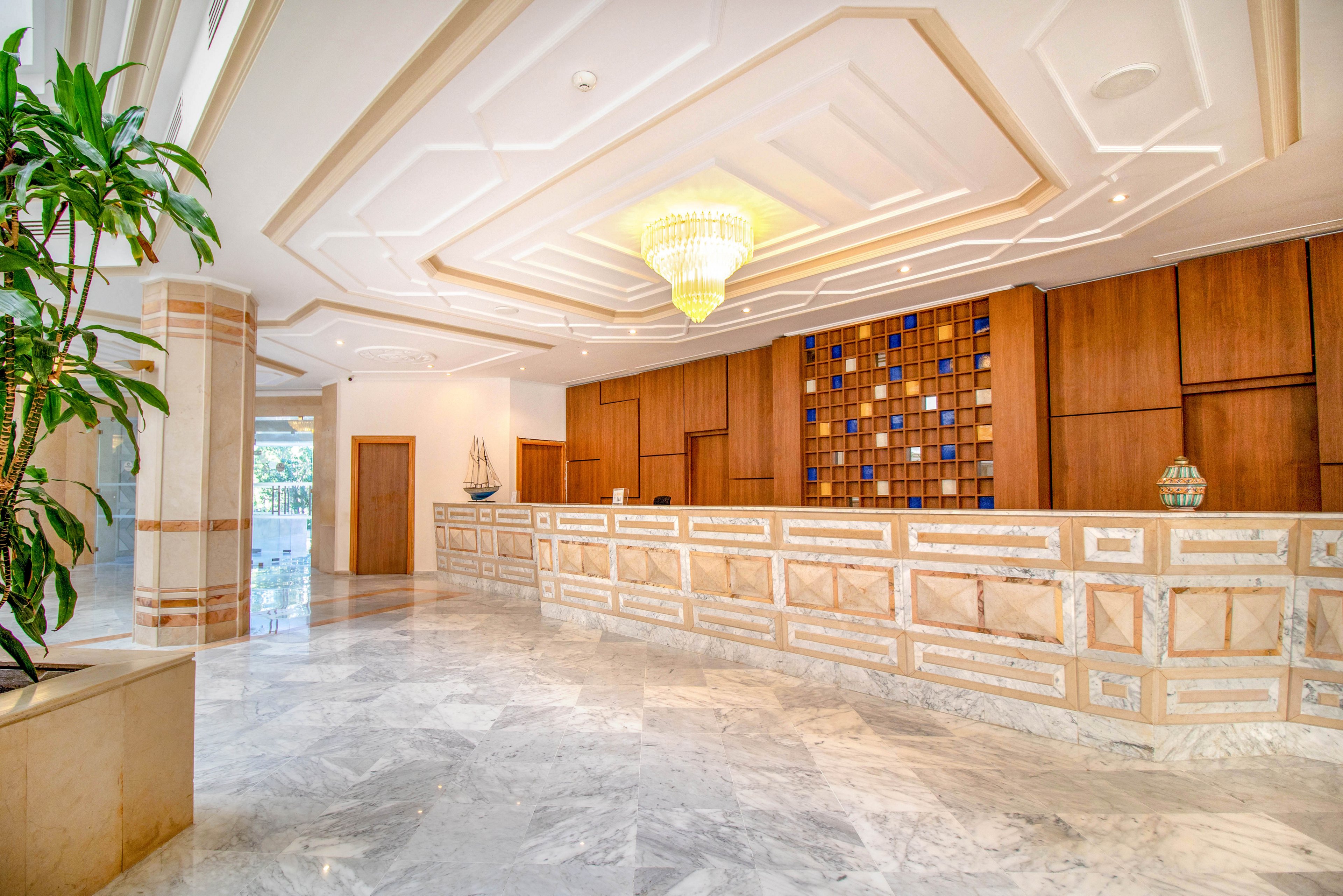 Spacious hotel lobby interior featuring a long marble reception desk as the central focus. The setting includes polished stone flooring, wood-paneled walls, and a decorative geometric wall behind the counter. A large chandelier hangs from the detailed ceiling, adding elegance and warm lighting to the space. Potted plants and decorative objects on the counter contribute to a welcoming, upscale atmosphere.