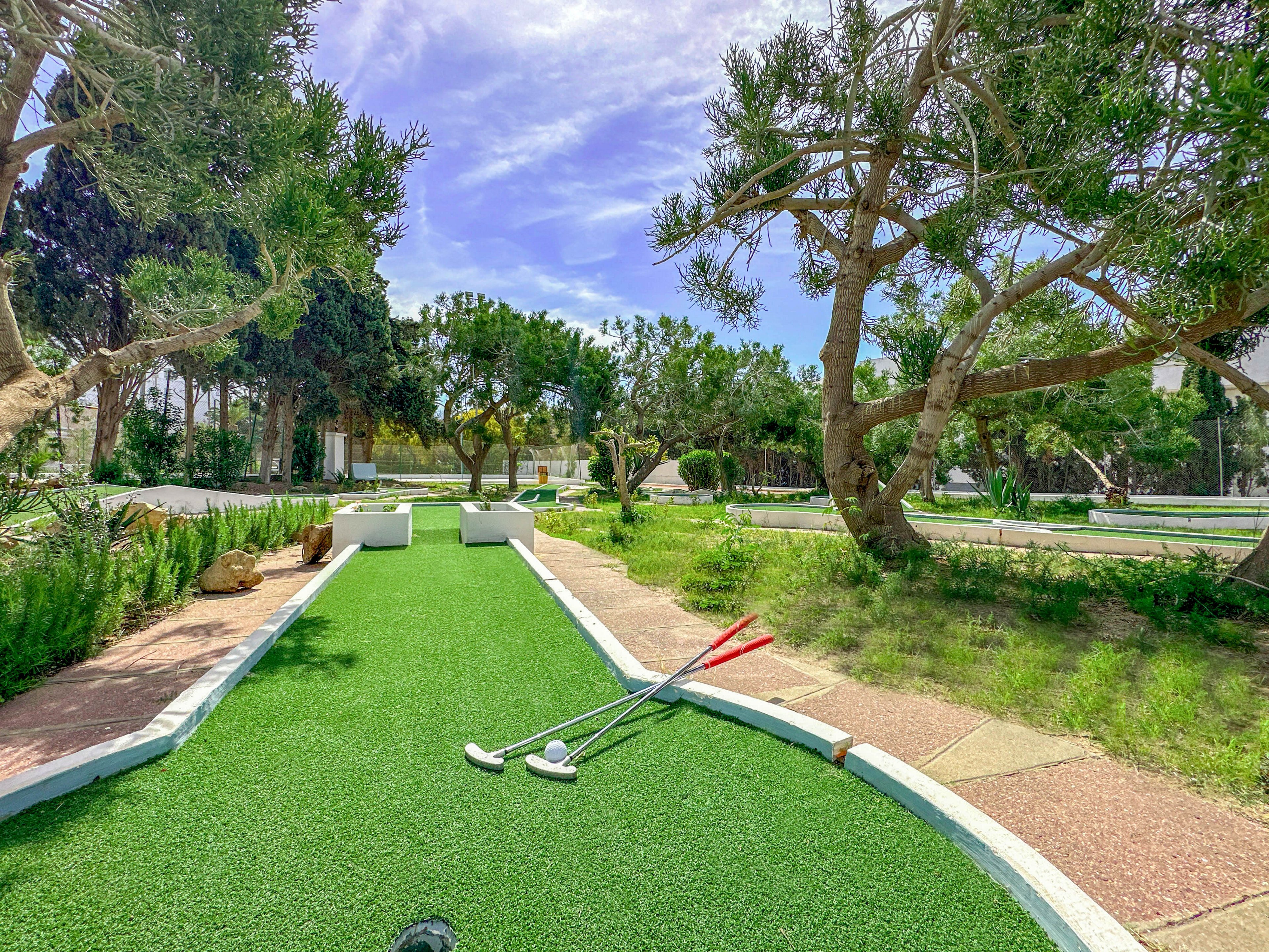 An outdoor mini golf course is shown under a bright, sunny sky. Several golf clubs lie on the artificial green near the start of a lane. Trees and shrubs surround the course, creating a relaxed park-like atmosphere. Pathways and low white borders define the playing area and guide the eye through the scene.