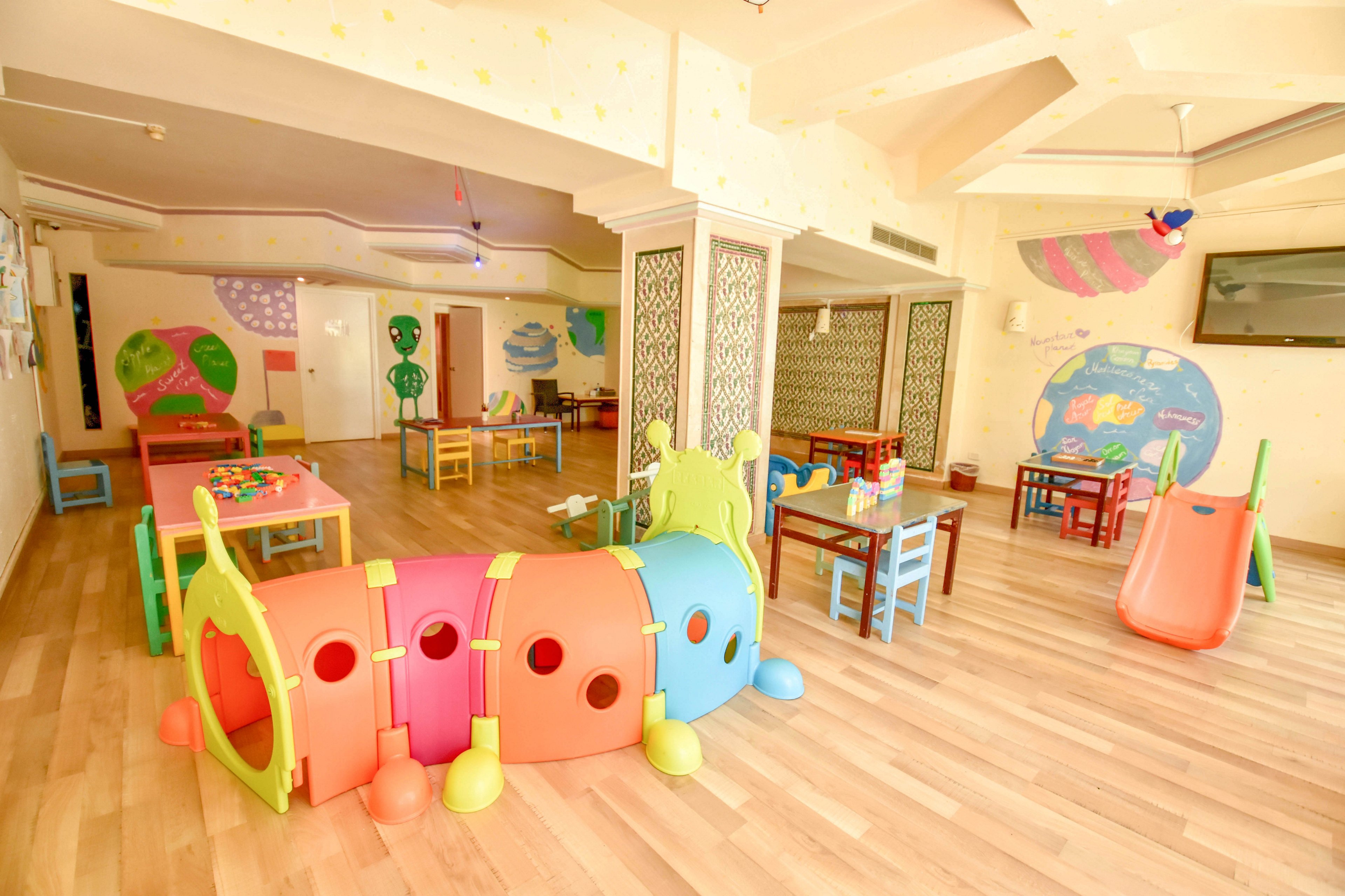 Colorful plastic play equipment fills a spacious indoor preschool playroom. A caterpillar tunnel, slide, and small tables with chairs are arranged on a light wooden floor. The walls are decorated with playful murals, shapes, and pastel colors, creating a cheerful environment. The room appears tidy and inviting, designed for young children’s activities and learning.