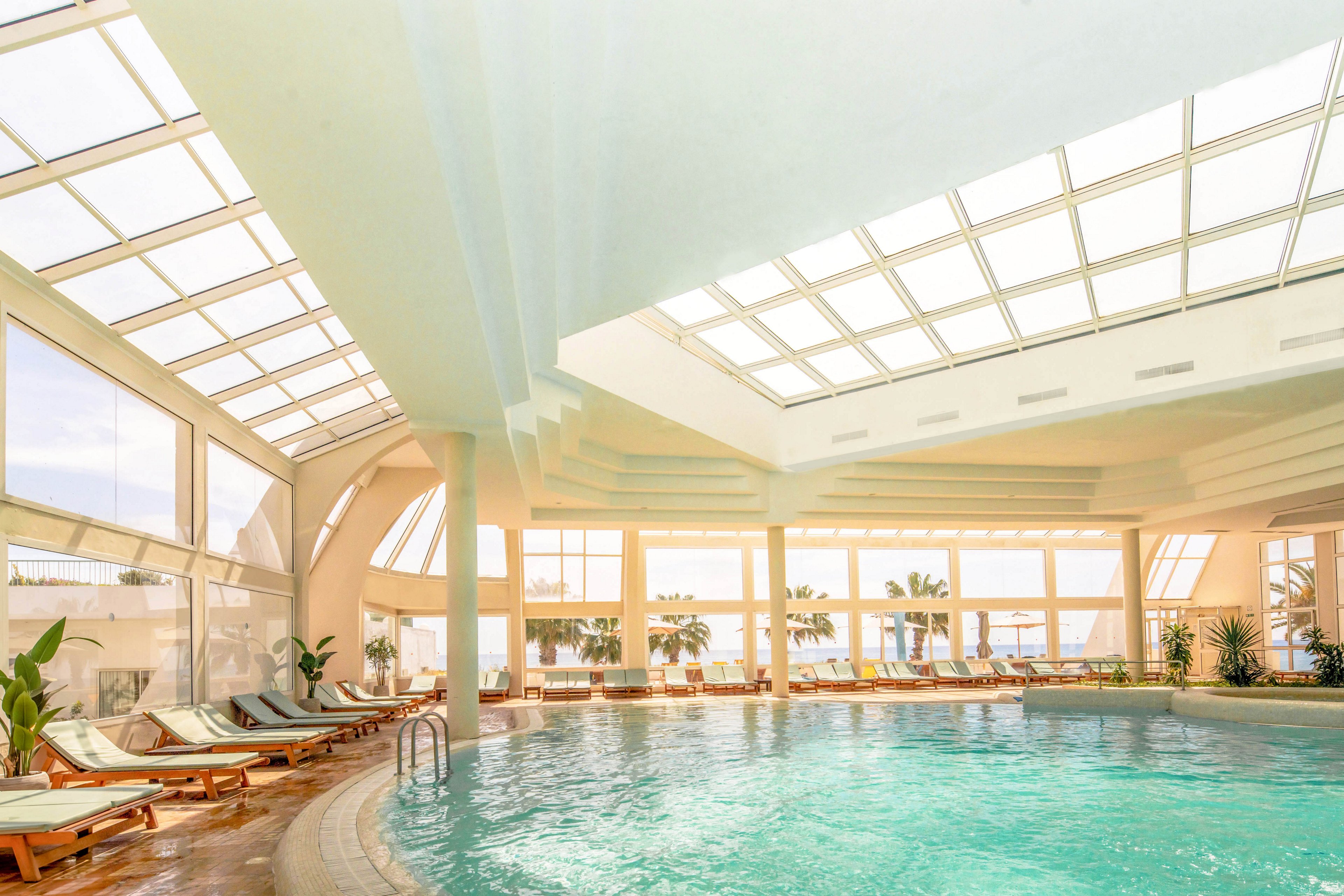 A spacious indoor swimming pool is surrounded by wooden loungers with cushions under a large glass ceiling. Sunlight floods the area through expansive windows, creating a warm and inviting atmosphere. Potted plants and views of palm trees outside add a tropical touch to the scene. The turquoise water and soft neutral tones emphasize relaxation and leisure in a modern resort setting.