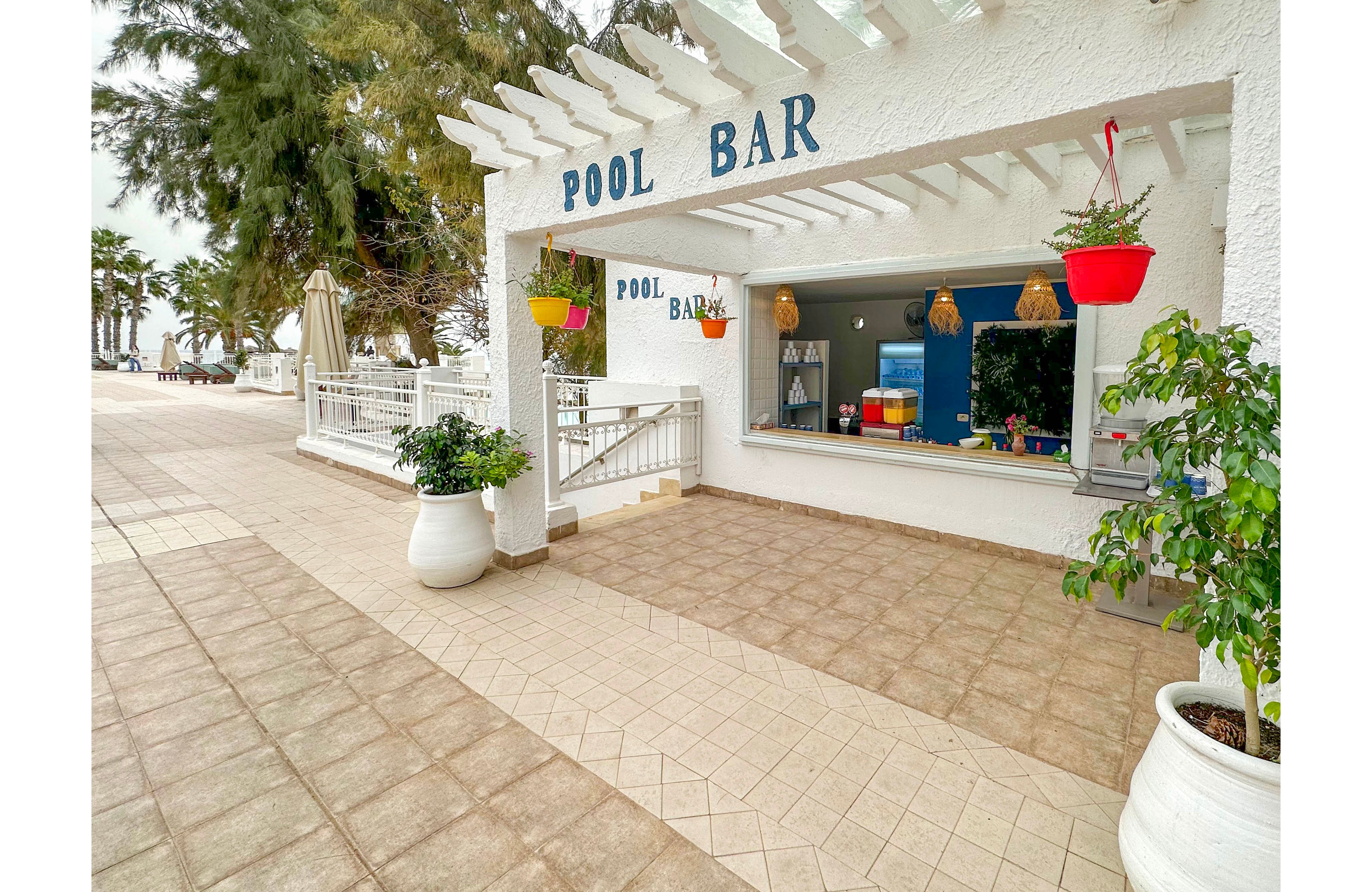 An open-air pool bar with a white pergola stands along a tiled seaside promenade. The bar counter window is open, revealing colorful items and hanging straw lamps inside. Potted plants and bright red hanging flower baskets add greenery and color to the clean, minimalist setting. Palm trees and lounge areas are visible in the background, suggesting a relaxed coastal resort atmosphere. The words "POOL BAR" appear prominently on the exterior wall.