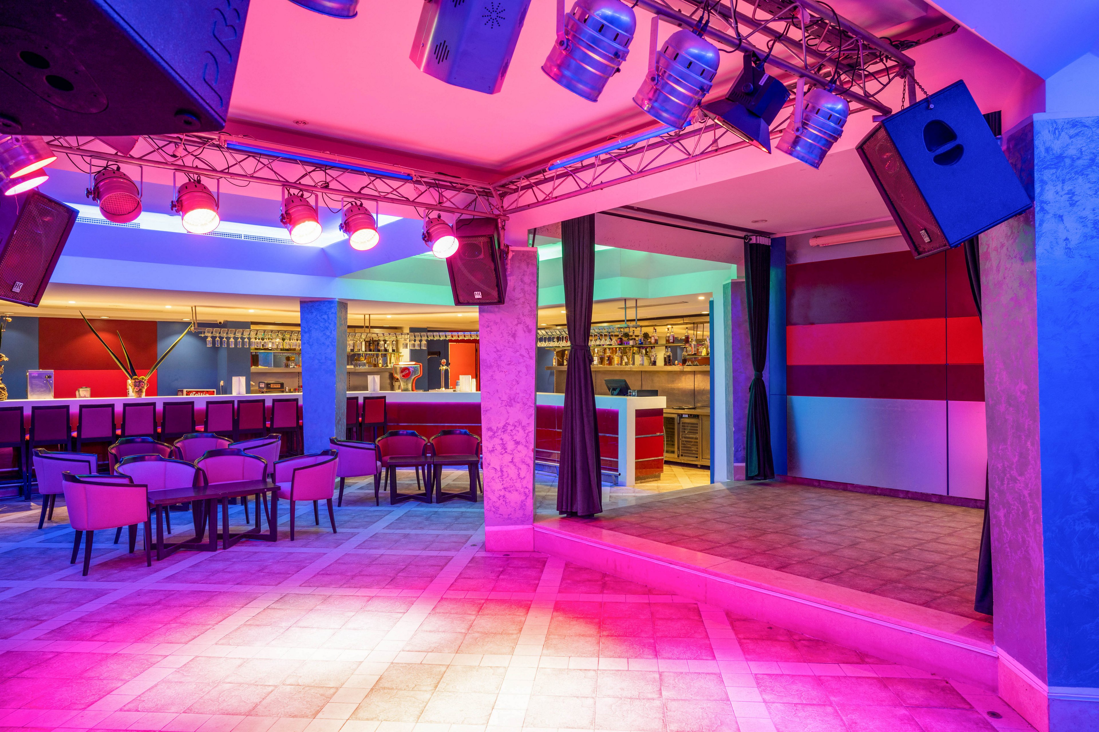 A vibrant nightclub interior features an empty stage lit by colorful spotlights. In the background, a long bar counter with stools and shelves of bottles is visible. Rows of chairs and small tables are arranged near the stage, suggesting a performance or dance area. The lighting casts pink, purple, and blue hues across the tiled floor and walls, creating a lively nightlife atmosphere.