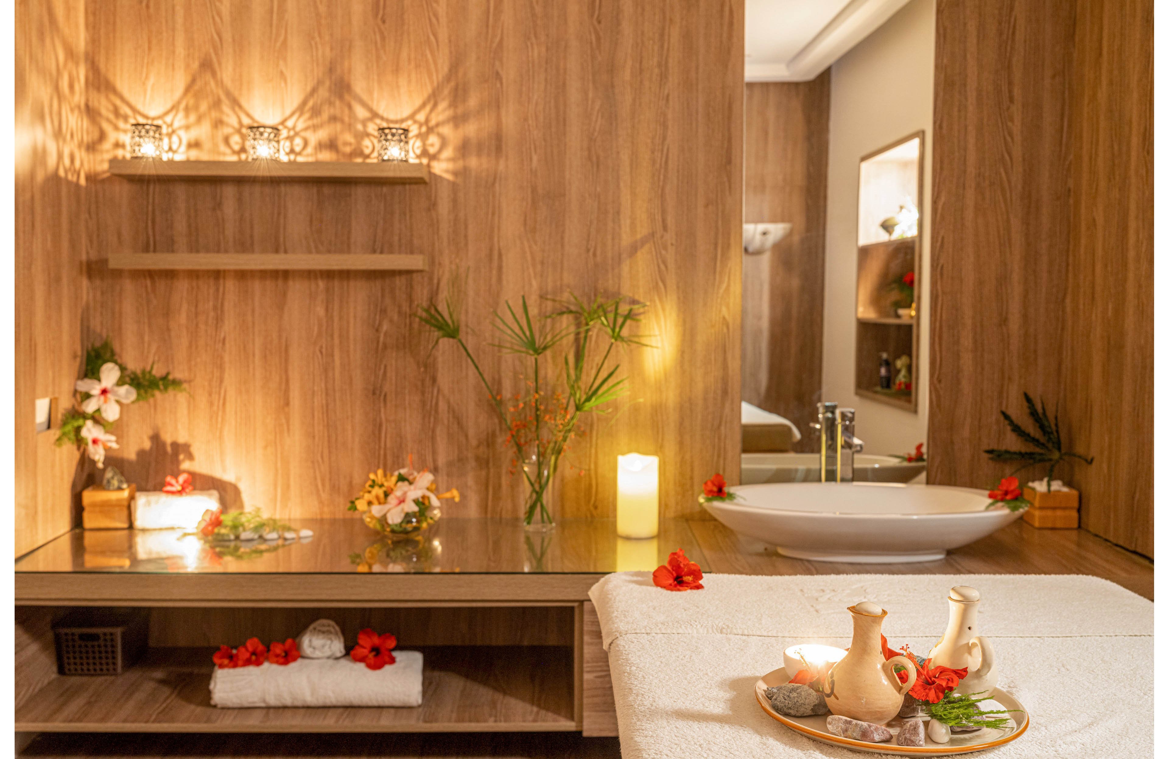 A serene spa treatment room features a massage table in the foreground with towels, flowers, and ceramic jugs arranged on a tray. A wooden vanity with a glass surface holds candles, decorative stones, and fresh flowers, creating a calming ambiance. A modern white basin sink and mirror are visible in the background, reflecting the soft lighting. The overall scene emphasizes relaxation, wellness, and a warm, inviting atmosphere.