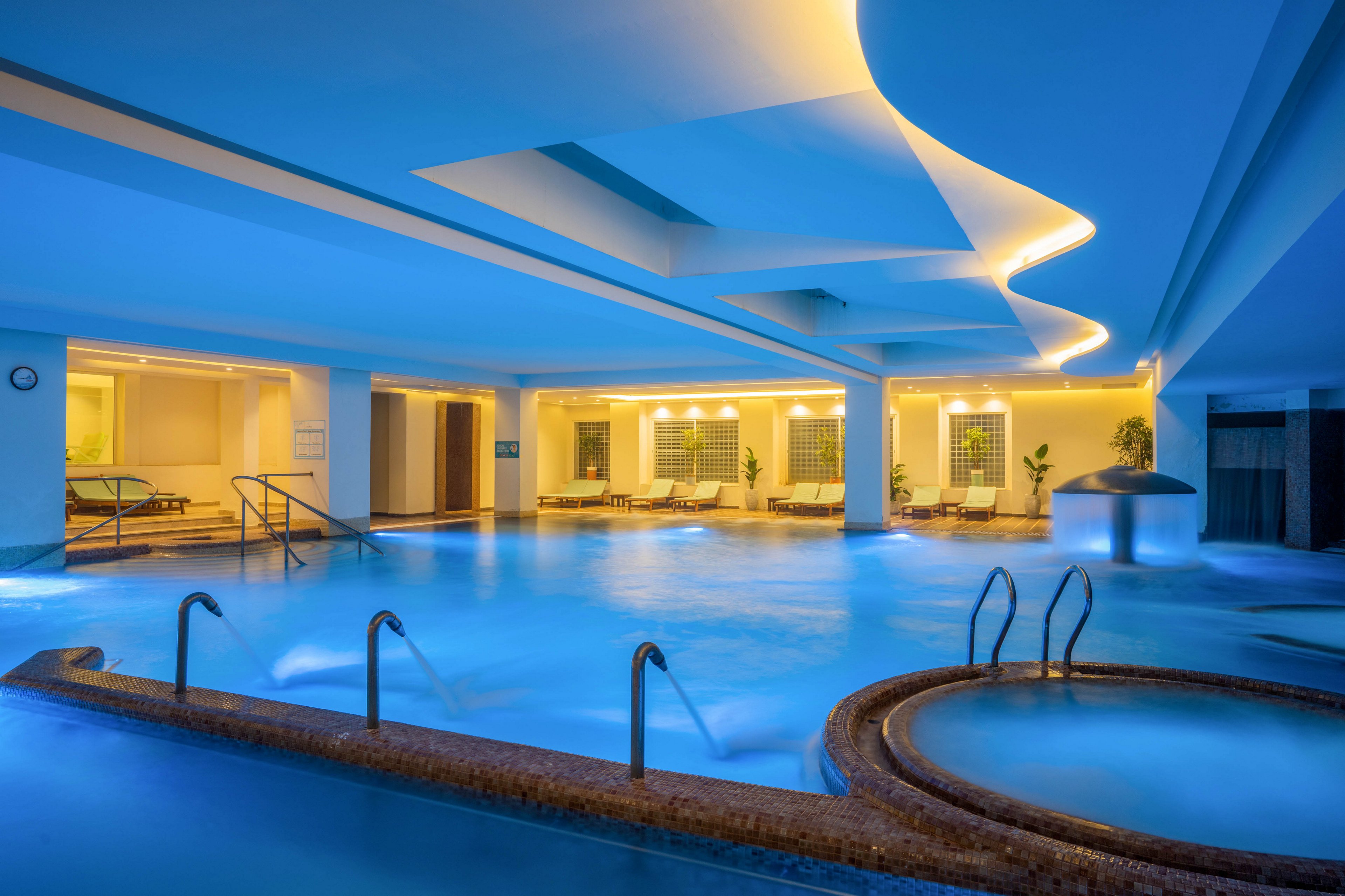 A contemporary indoor spa pool glows with soft blue lighting, creating a tranquil atmosphere. Multiple pools and curved hot tub areas are surrounded by smooth stone edges and stainless steel handrails. Warm yellow accent lights highlight the ceiling design and the seating area with lounge chairs in the background. Potted plants and clean architectural lines contribute to a luxurious wellness and relaxation setting.