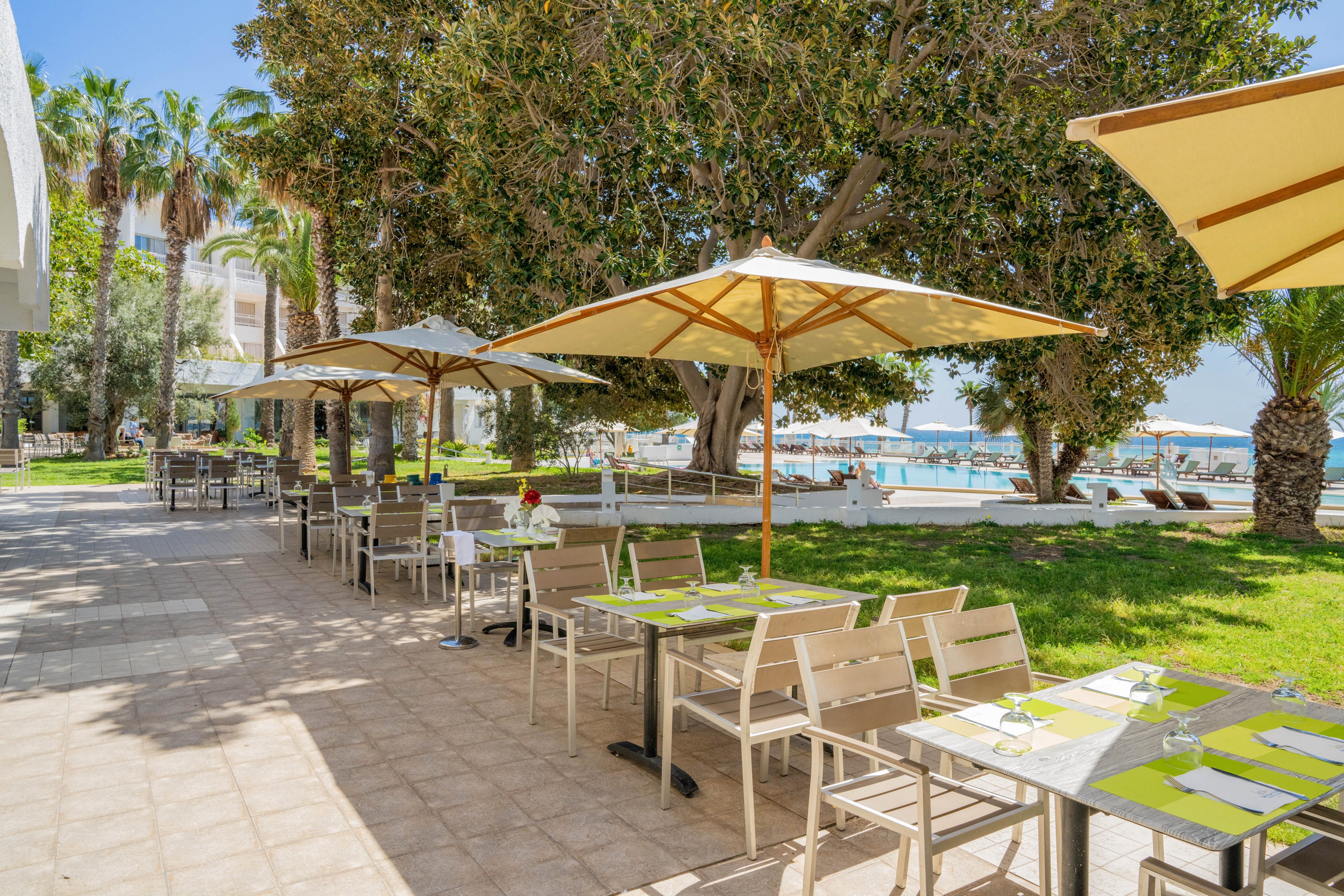 An outdoor restaurant terrace is set with tables and chairs under large beige umbrellas. The scene is bright and sunny, with green grass and tall palm trees providing a resort-like atmosphere. In the background, the blue sea and a marina with boats are visible, suggesting a coastal location. The overall setting feels relaxed and inviting for al fresco dining.