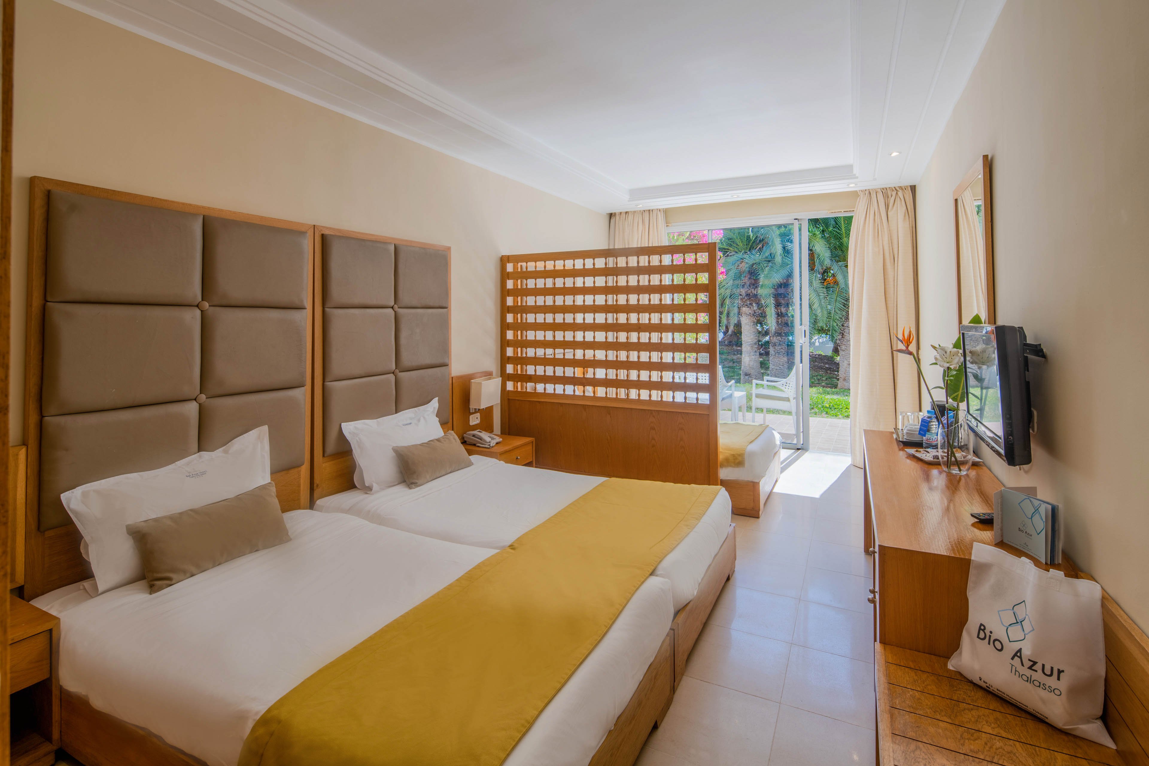 A modern hotel room features two neatly made twin beds with mustard yellow runners and padded headboards. A wooden partition separates the sleeping area from a small sitting space with a daybed near the balcony. Natural light floods in through large glass doors that open to a garden with palm trees. Neutral beige walls and light tile flooring create a calm, contemporary atmosphere, complemented by a desk, television, and decorative flower.
