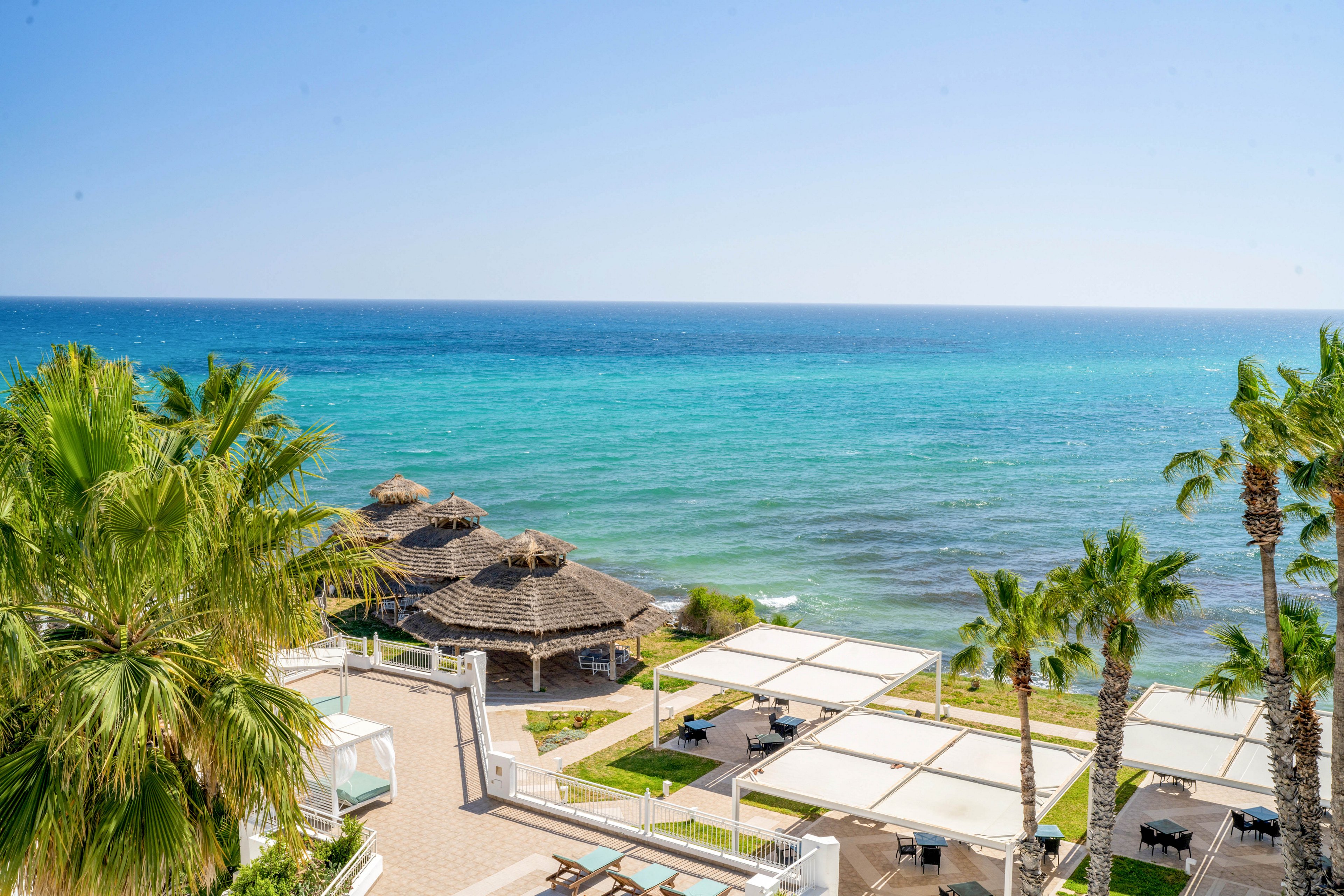 A bright seaside resort overlooks a calm turquoise ocean under a clear blue sky. Palm trees frame the scene, adding a tropical feel to the coastal landscape. Thatched-roof structures and white pergolas provide shaded seating areas near the water. The overall atmosphere is relaxed and inviting, suggesting a warm vacation destination.